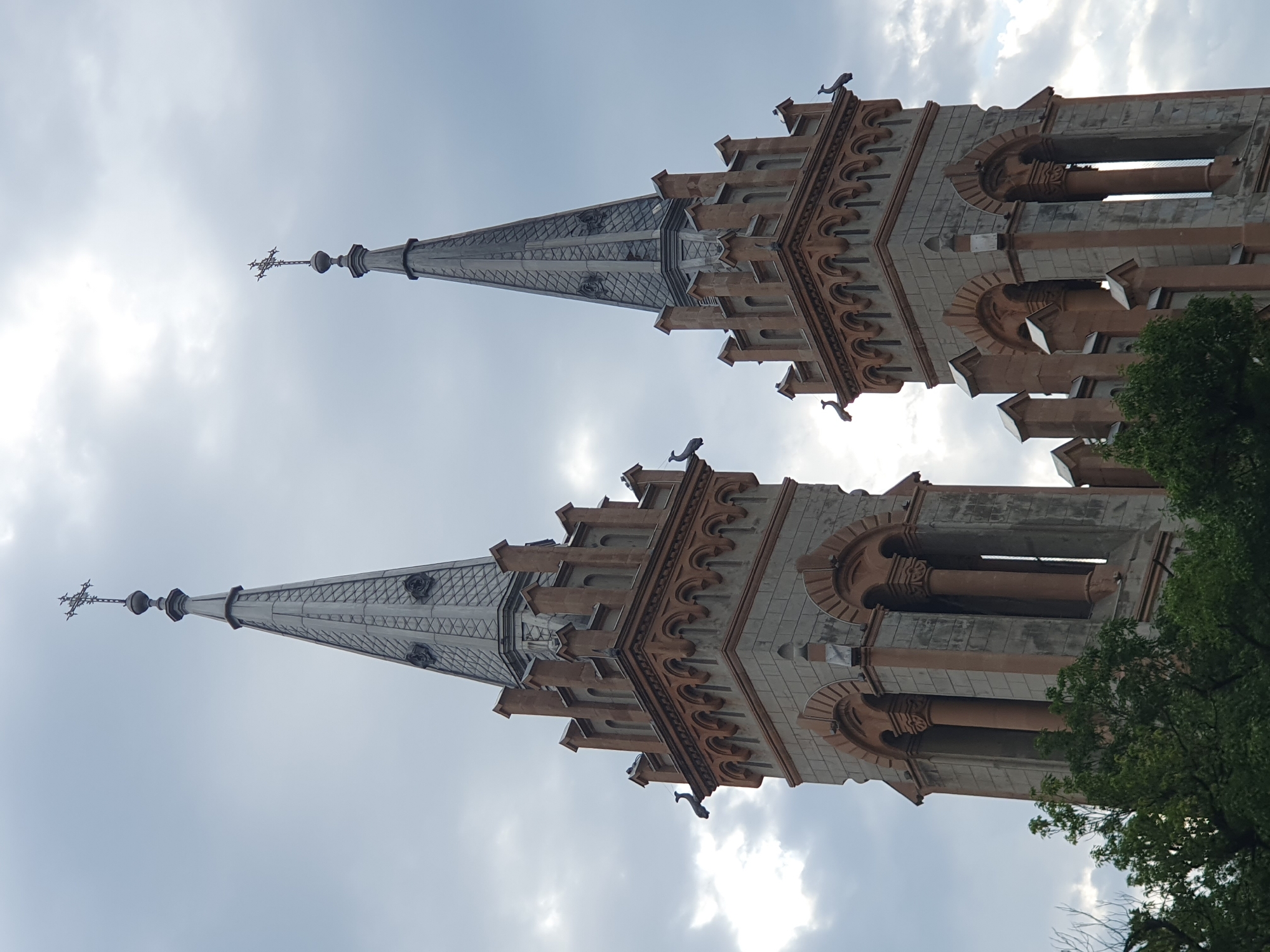 The image displays the towers of the Holy Trinity Church in Batumi, Georgia, against a cloudy sky. The church features two prominent spires, each topped with a cross. The towers are constructed with a combination of grey stone and reddish-brown architectural elements. Gargoyle-like figures are visible protruding from the upper sections of the towers, and decorative arches and columns adorn the structures. Lush green trees are visible at the base of the towers. The overall scene is dominated by the imposing architecture of the church and the dramatic, overcast sky.
