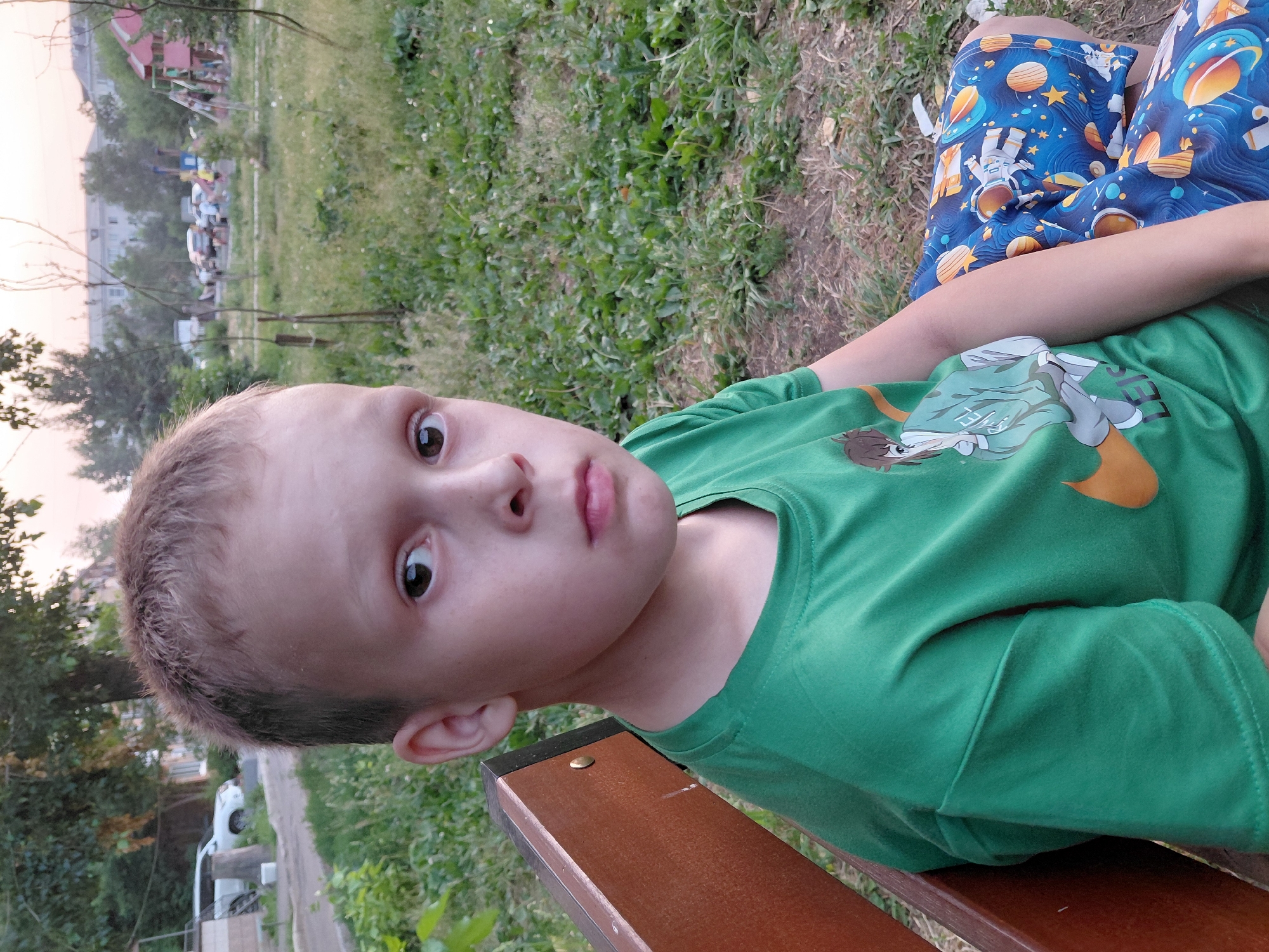 The media file shows a close-up, vertically oriented shot of a young boy sitting on a wooden bench outdoors. He is wearing a green t-shirt with a cartoon character print on it and blue shorts with a space-themed pattern featuring astronauts and planets. The boy is looking towards the camera with a neutral expression.
In the background, there is a grassy area with some scattered leaves, and beyond that, a playground with some structures and parked cars are visible. Trees and buildings are also present in the distance, suggesting an urban or suburban park setting. The lighting suggests it is either early morning or late afternoon. The weather appears to be mild and clear.
No visible text is present in the media file. The scene is likely a casual outing at a park in Oskemen, Kazakhstan.