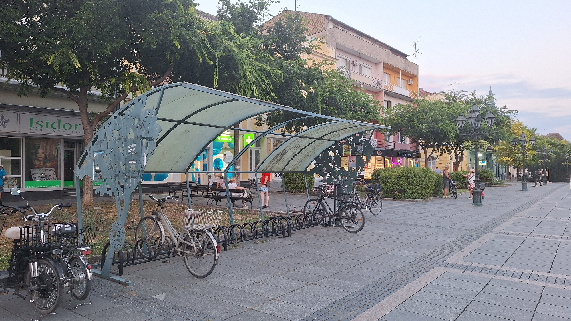 The image depicts a street scene in Sombor, Serbia, likely during late afternoon or early evening. In the foreground, a row of bicycles is parked under a covered structure, suggesting a public space or park. The bicycles are of various types, including what appears to be an electric bicycle on the far left. The covering is translucent, allowing diffused light to pass through.
Behind the bicycle racks, benches are visible, and a few individuals are seated, engaged in conversation or simply resting. A store with a prominent sign reading "Isidor PRO COSMETICS" is visible to the left, indicating a commercial area. The architecture of the buildings lining the street suggests a European urban setting, with multi-story structures featuring balconies and shops on the ground floor.
The street itself is paved with stone tiles, and decorative lampposts line the walkway, some featuring multiple lights. Lush green trees are interspersed along the street, providing shade and contributing to the pleasant atmosphere. The sky appears overcast or nearing dusk, with soft lighting casting a warm glow on the scene.
Further down the street, more people can be seen walking and interacting, and other shops and businesses are identifiable by their signage and illuminated displays. The overall impression is one of a lively, pedestrian-friendly urban environment.