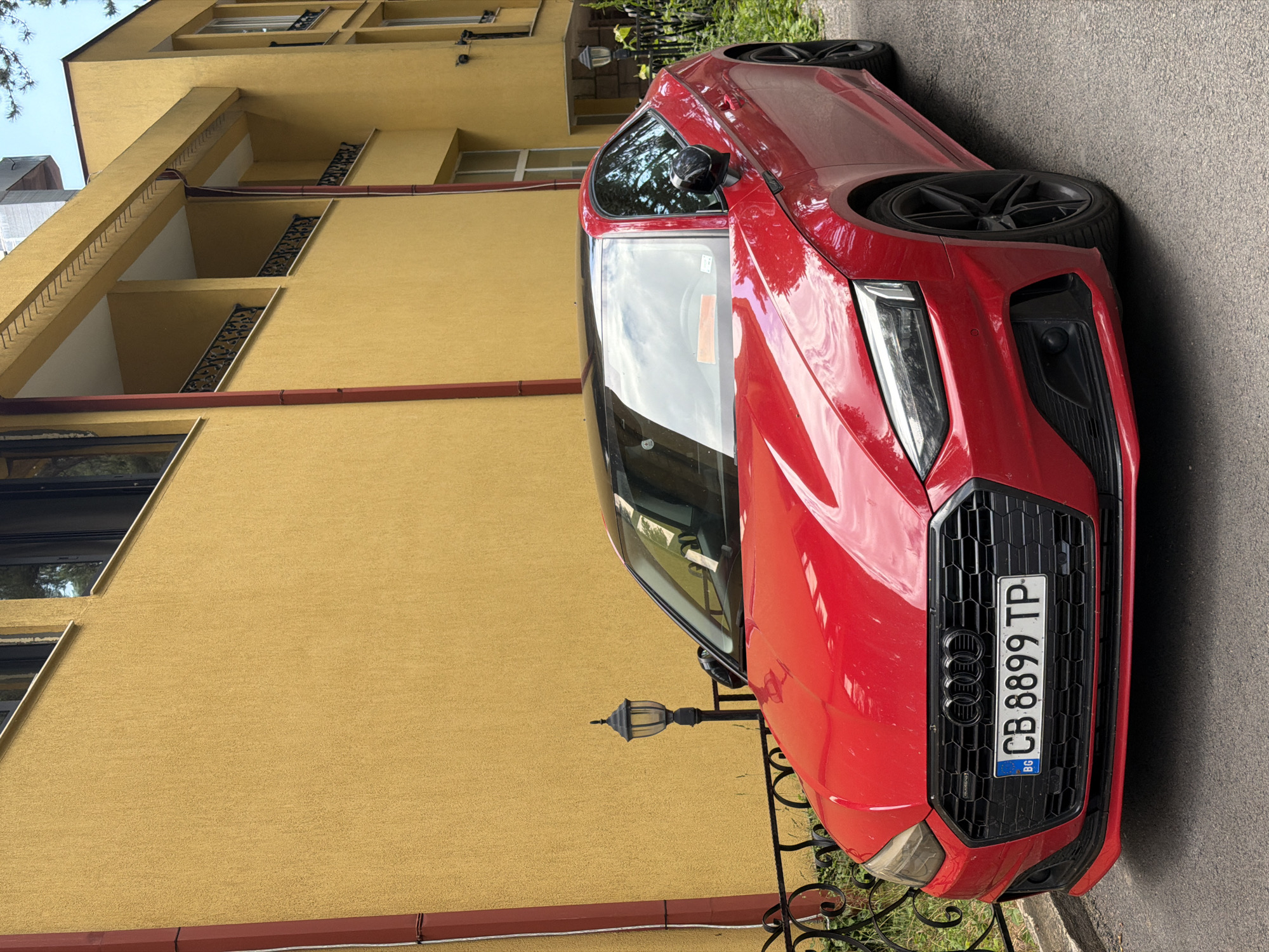 A red 2020-2024 Audi A5 Coupe is centrally parked on an asphalt surface, angled slightly towards the right. The vehicle features a gloss black roof, black multi-spoke alloy wheels, and a black honeycomb grille displaying the Audi rings logo and a "Quattro" badge. A Bulgarian license plate, "BG CB 8899 TP," is affixed to the front bumper. The car's body reflects bright daylight, showing sky and surrounding structures.
Behind the vehicle stands a multi-story building with a textured, mustard-yellow stucco facade. Red vertical downspouts or pipes are visible on the building's exterior, notably on the left and near the center. The building incorporates recessed balconies on its upper floors, each featuring ornate black wrought iron railings. Dark-framed windows are present across different levels, some exhibiting reflections.
To the left of the Audi, a black wrought iron fence with decorative elements separates the pavement from a small area of green foliage. A black vintage-style street lamp with a glass lantern is positioned behind this fence, near the vehicle's hood. Another similar lamp is partially visible on the right side of the image, behind the car's rear quarter panel, amidst green plants. The pavement is dark gray asphalt. No individuals are depicted in the scene. The image is brightly lit, suggesting daytime conditions. The setting is identified as the city of Kyustendil, Bulgaria.