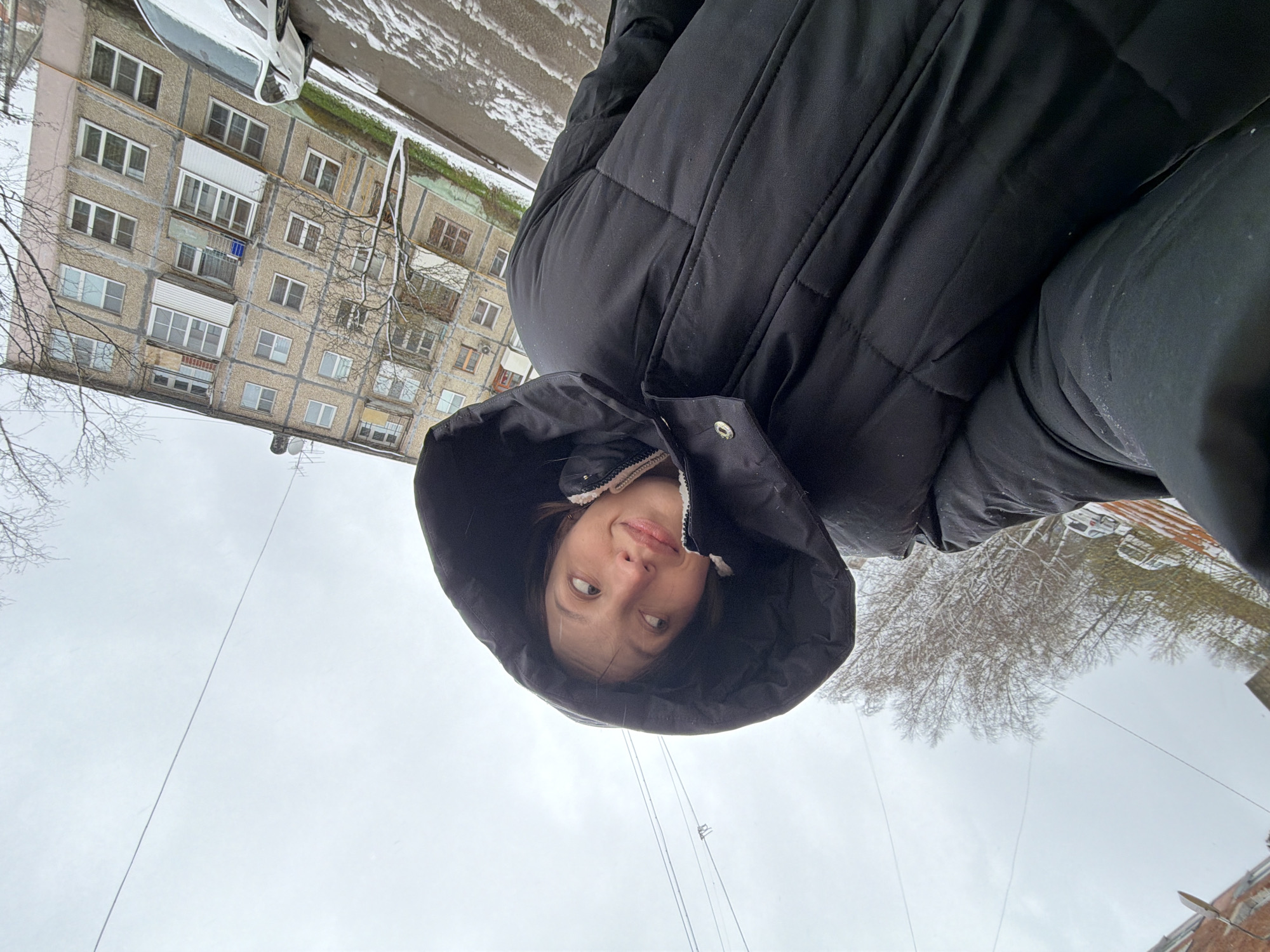 The image captures a low-angle outdoor scene on an overcast, snowy day in Reshetnikovo, Russia. In the foreground, a person, likely a young woman, looks upwards towards the camera with a slight, playful expression. She is wearing a dark, possibly black, puffer jacket with the hood pulled up, framing her face. Her dark hair is visible beneath the hood, and she has a fair complexion.
The background features a multi-story apartment building constructed from concrete, characterized by numerous windows and balconies. The architectural style is consistent with residential buildings found in Russia. Snow is visible on the ground and covering parts of the building ledges, indicating cold weather conditions. A white car is parked on the snowy ground near the building. Bare trees, stripped of their leaves, are scattered throughout the mid-ground, further suggesting winter or early spring. Overhead, a uniform grey sky dominates, with thin power lines or cables traversing the frame. No discernible text is visible in the image. The overall impression is a candid shot taken during an overcast, snowy day in a residential urban environment.
