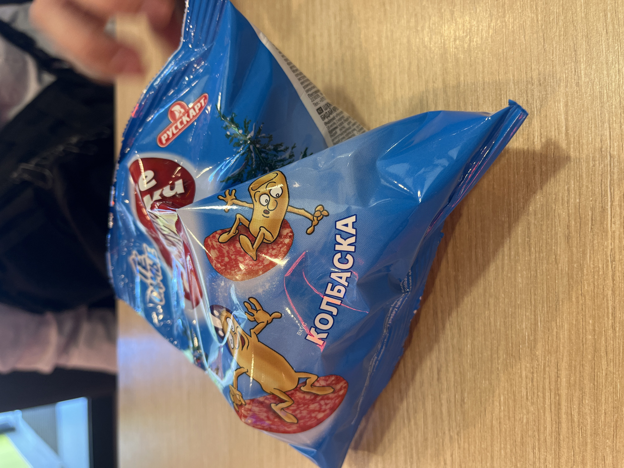A bright blue, crinkled plastic bag of what appears to be sausage snacks rests on a light brown wooden table. The bag features cartoonish illustrations of a yellow, sausage-like character with wide eyes and arms, depicted as if surfing on slices of salami. The prominent text on the bag is "КОЛБАСКА," which translates to "Sausage" in Russian. Above this, a red circular logo with white text is partially visible, as is the word "РУССКАРТ" within a red oval to the right. In the background, out of focus, a person's arm and shoulder are visible, wearing a dark jacket over a light-colored shirt, and another person's blurred form. The setting is indoors, likely a cafe or restaurant, given the table and the presence of people. There is no specific indicator of the city of Bryansk, Russia, in the image itself.