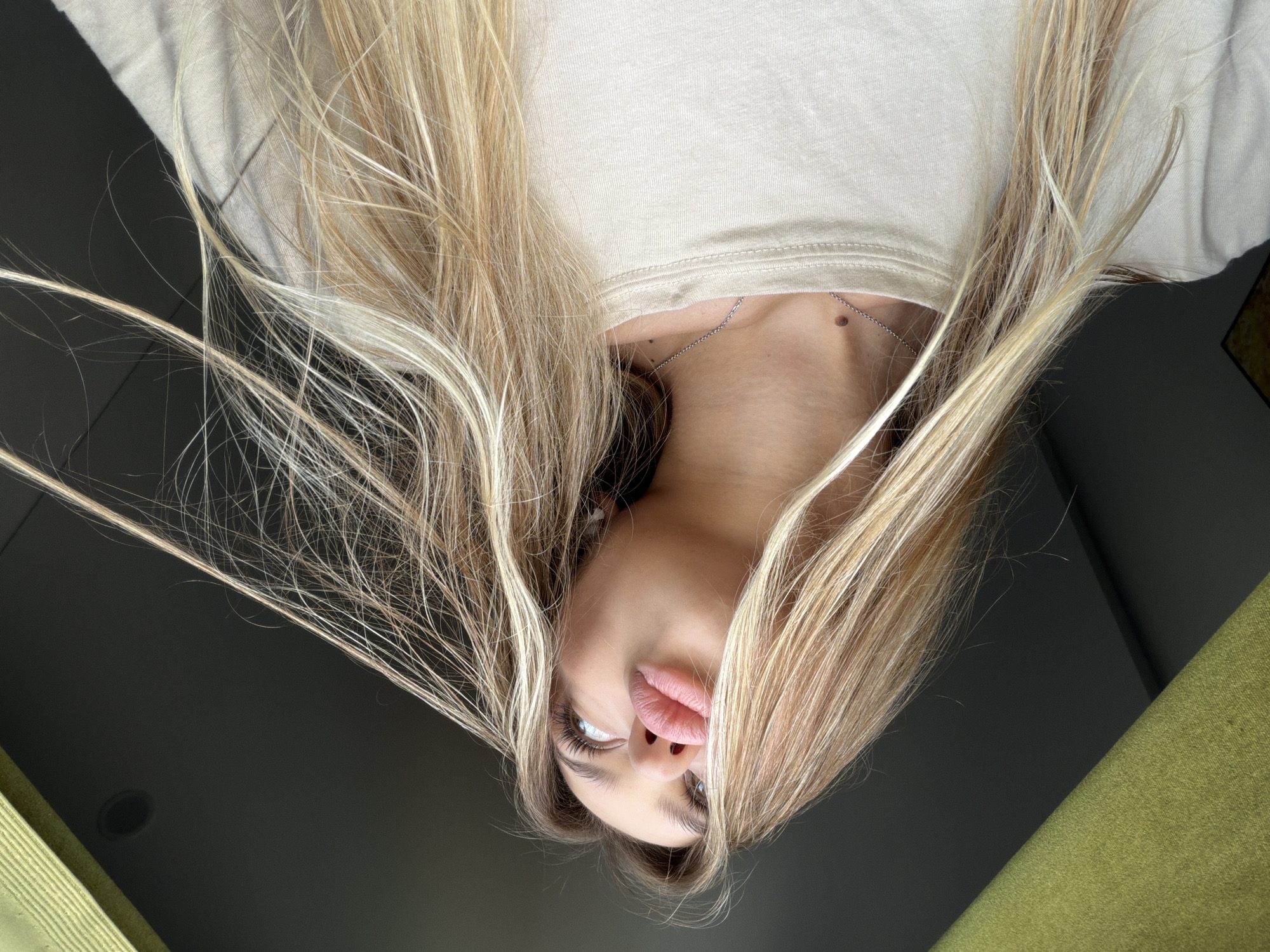 A low-angle, close-up shot shows a young woman with long, blonde hair styled with loose waves. She is wearing a light-colored t-shirt and a thin silver necklace. Her lips are puckered as if blowing a kiss, and her eyes are looking upwards. The background is dark, with a diagonal line separating it from a textured, lime green surface in the upper left corner. No identifiable landmarks or specific environmental details suggest a location in Đà Nẵng, Vietnam.