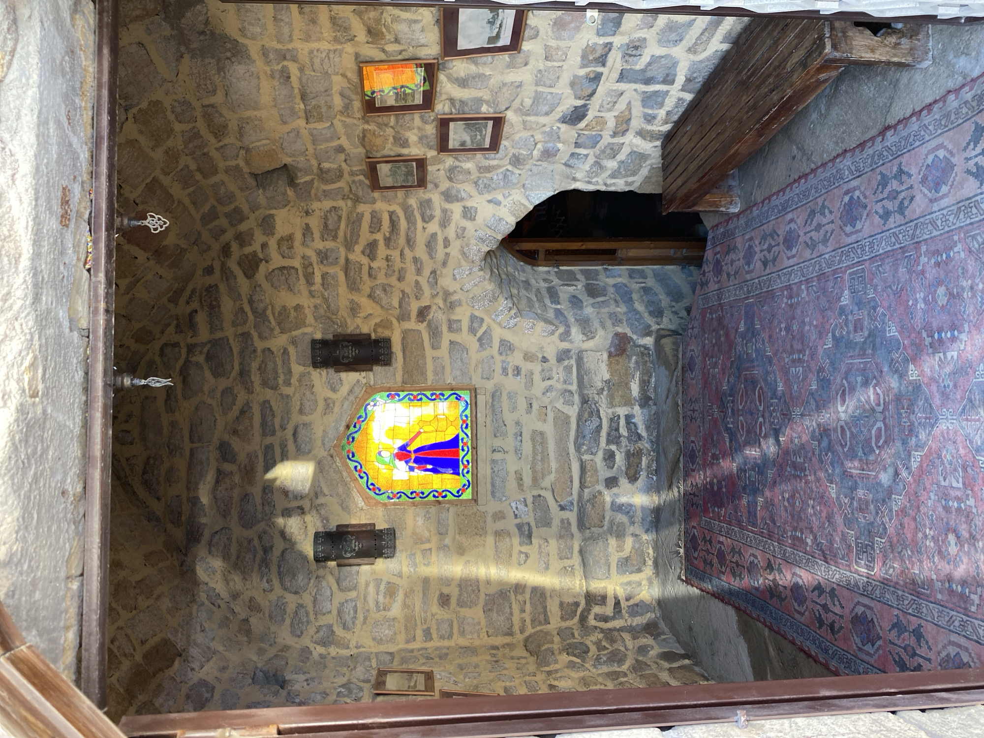 The image shows the interior of a stone-walled room, possibly part of a historical building in Derbent, Russia. The room features a vaulted stone ceiling and rough-hewn stone walls. A large, patterned rug covers the floor in the foreground. To the left, there are two framed pictures on the wall. In the center of the back wall is a small, arched stained-glass window depicting two figures, one in blue and red. To the right of the stained-glass window are three more framed pictures, and to the far right, a wooden bench. An arched doorway with a wooden door is visible in the far right wall. Light streams into the room from the stained-glass window, casting shadows on the floor and walls.