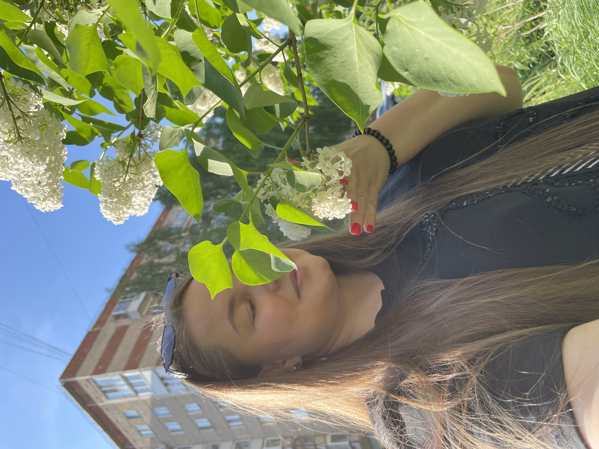 The image depicts a woman lying on the grass, possibly in a park or green space in Izhevsk, Russia. Her face is tilted towards the camera, eyes closed as if in relaxation. She is wearing a black top and has long, light brown hair spread around her. A branch of a lilac bush, with white blossoms and green leaves, is positioned above her head. Her hand, adorned with red nail polish and a bracelet, lightly touches the flowers.
In the background, a building can be seen, suggesting an urban location within Izhevsk. The sky is blue, and the sunlight illuminates the scene.