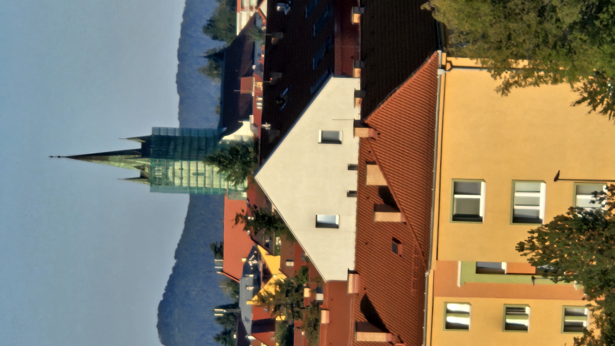 The image is an aerial view of a city skyline in Prešov, Slovakia, dominated by a church tower undergoing renovation, with scaffolding covered in green netting. The church tower is tall and slender, with a pointed spire topped by a cross, and features Gothic architectural elements. Below the church, a dense urban landscape unfolds, characterized by a variety of buildings with terracotta-tiled roofs, painted facades in warm colors like yellow and orange, and white-framed windows. The foreground features apartment buildings with multiple stories and numerous windows. Lush green trees are scattered throughout the scene, some in the immediate foreground and others nestled between the buildings and in the mid-ground. In the background, rolling hills form a natural backdrop to the urban environment. The sky is a clear, light blue, suggesting fair weather. The image captures a mix of architectural styles and the natural integration of greenery within the city.