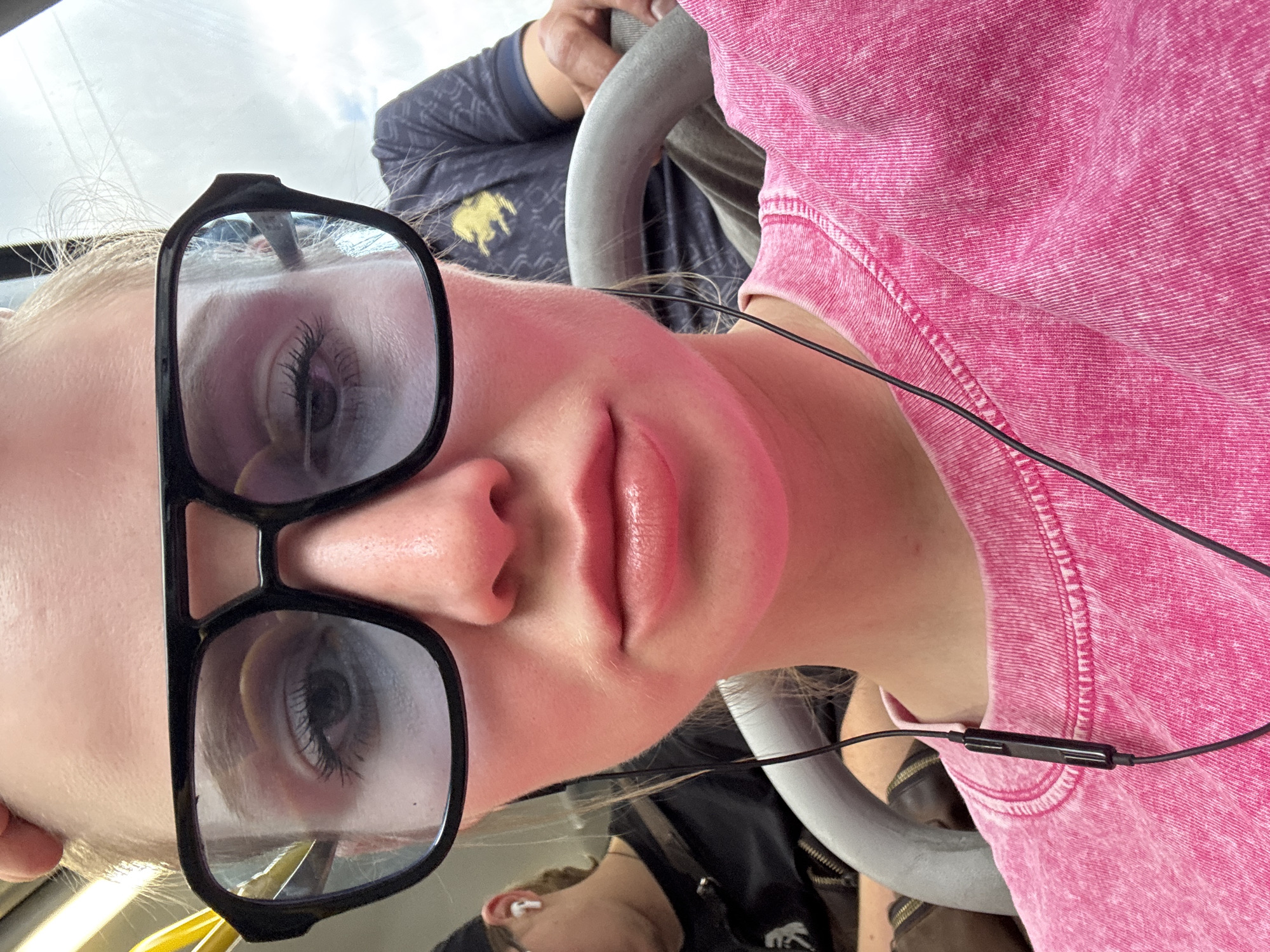 A close-up portrait captures a young person, likely female, with blonde hair, seen from a slightly low angle. She wears large, black-framed glasses through which her eyes, accentuated by long eyelashes, are visible. Her lips are a soft pink and slightly parted. She is dressed in a vibrant pink, long-sleeved top with a distinct textured or acid-washed appearance. A black wired earphone cable, featuring a small control module, extends from her ear area across her shirt.
The setting appears to be public transport, possibly a bus or train, suggested by a grey, curved handrail or support bar. In the background, another person is partially visible, wearing a dark blue top adorned with a yellow emblem resembling a polo player. Reflections in the subject's glasses show a cloudy sky, indicating daytime, and a partial view of another individual wearing an earbud. The overall impression is a casual moment during travel in Saint Petersburg, Russia. No visible text is present other than the emblem on the background clothing.