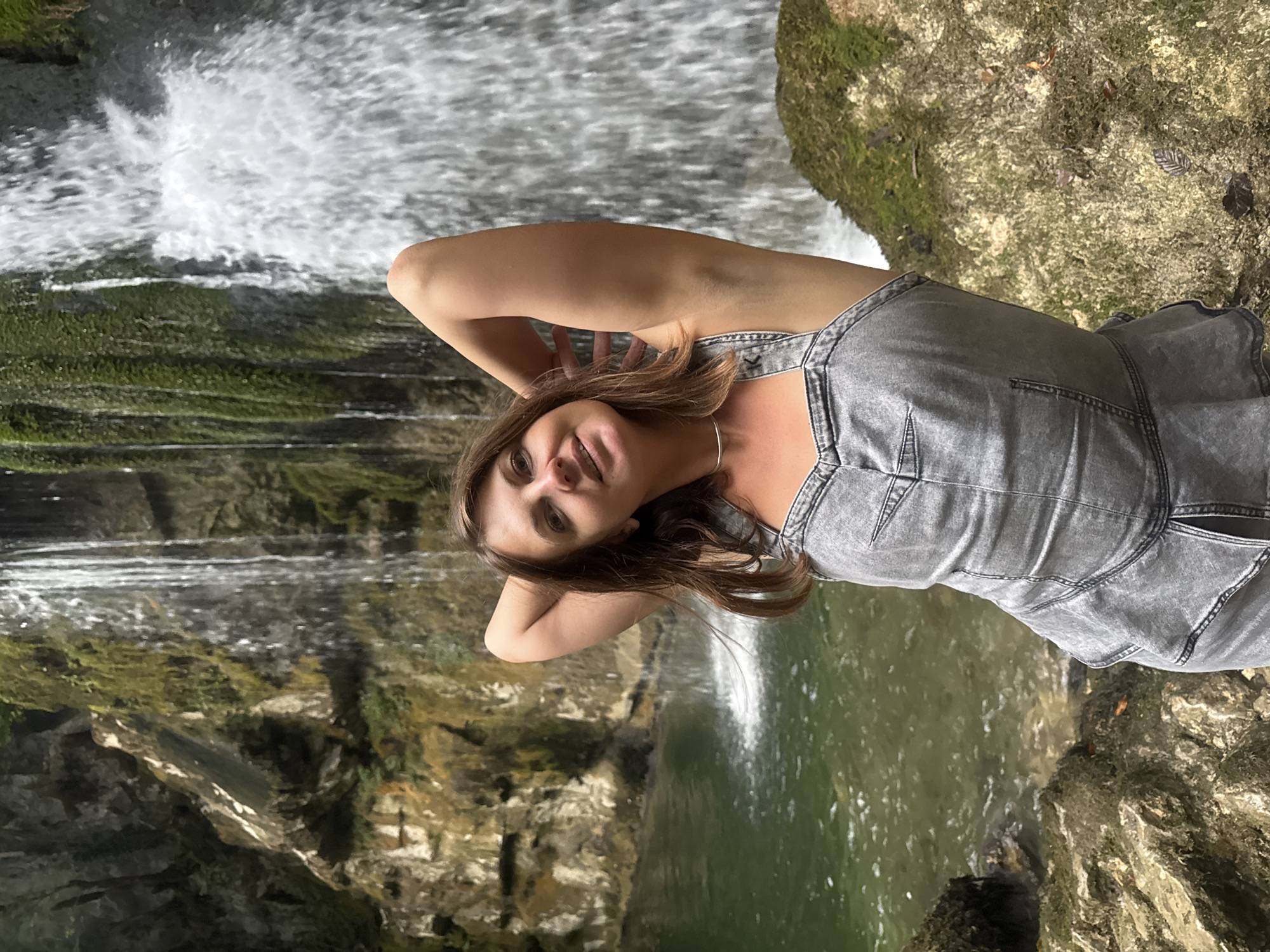 A medium shot captures a woman posing with her arms behind her head in front of a waterfall. She has brown hair and is wearing a grey denim outfit. The waterfall cascades down a rocky, moss-covered cliff into a clear green pool. The background suggests a natural, lush environment. The location is identified as being in or near Saint-Claude, France.