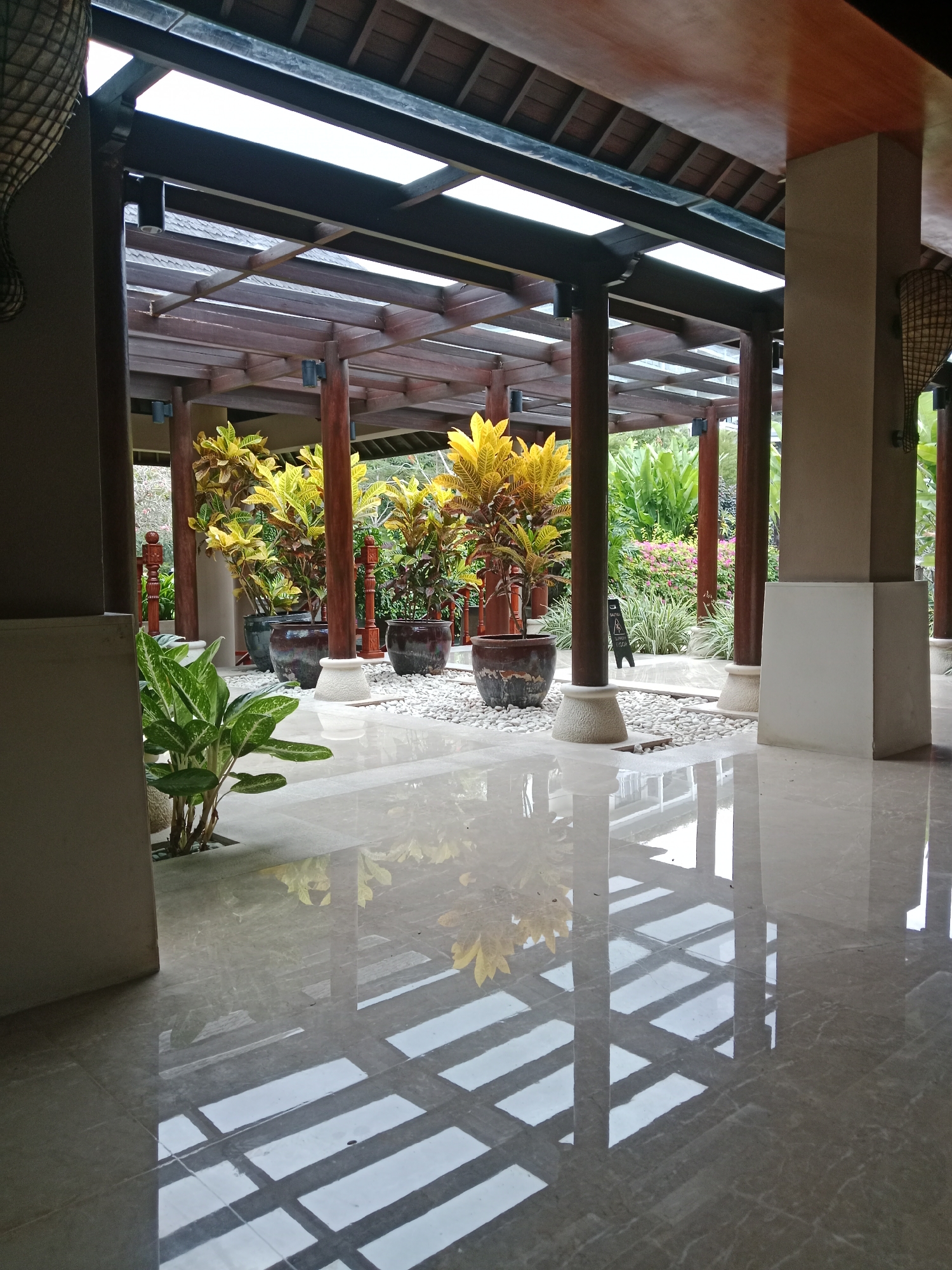 This is a photograph of a covered walkway or patio area at a resort in Kuta Selatan, Indonesia. The architecture features a wooden beamed roof with some translucent panels, supported by large, dark wooden pillars. In the center of the walkway, there are several potted plants with yellow and green foliage. One large green plant with broad leaves is situated in the foreground to the left. The floor is highly polished, reflecting the overhead structure and the plants. The background reveals a lush garden area with more greenery and some pink flowers. There are no people visible in the image. The scene conveys a sense of tranquility and tropical ambiance.