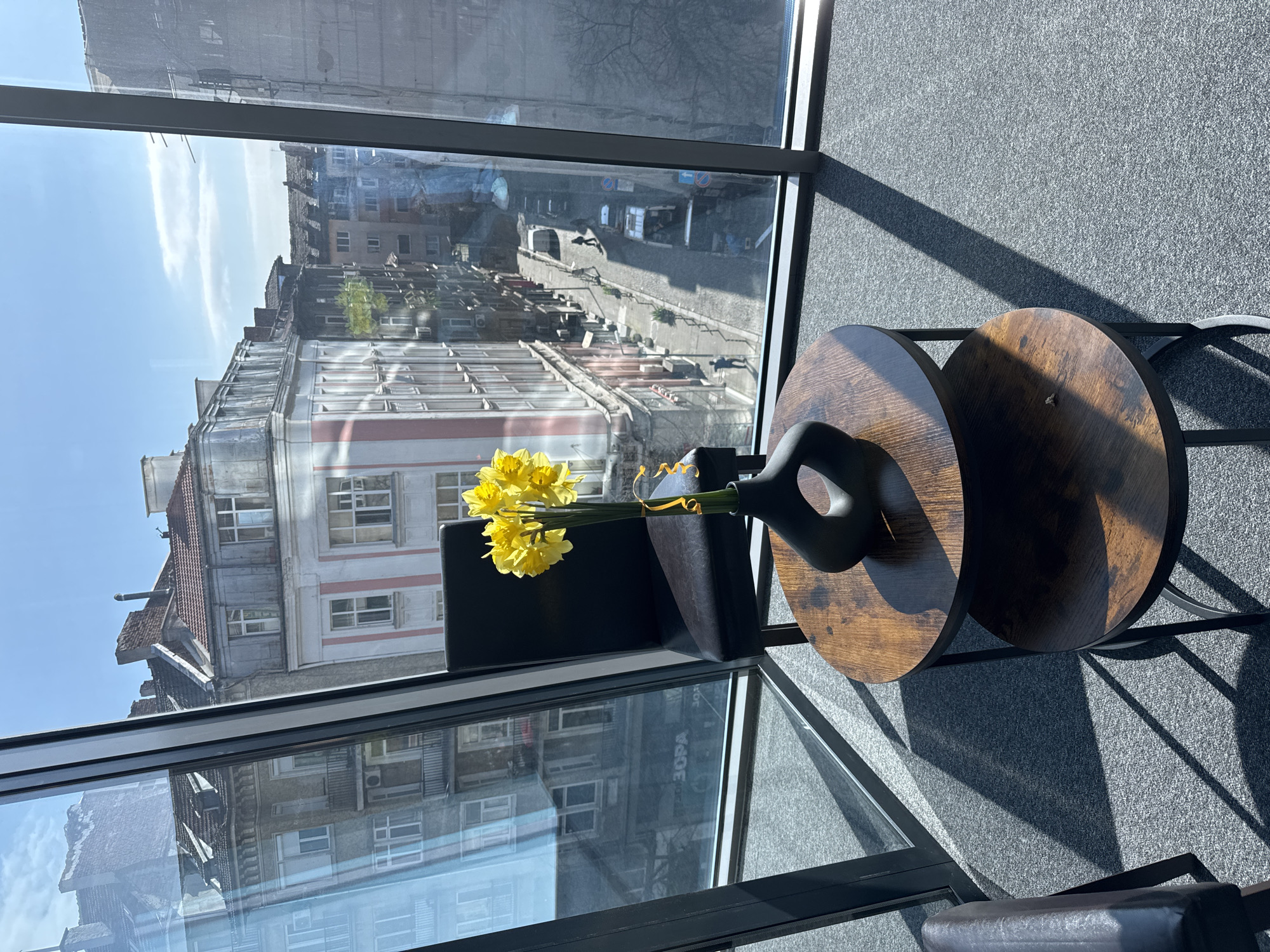 A high-angle indoor shot depicts a window view in what is likely Sofia, Bulgaria, as evidenced by the architecture visible through the glass. The scene includes a pair of circular wooden tables supporting a black vase filled with yellow daffodils. A black cushion or small chair is positioned near the window. The view through the window reveals a street lined with buildings exhibiting a mix of architectural styles. The weather appears to be clear, with sunlight casting shadows on the interior floor and walls. The flooring is a medium-gray, textured material. The window has a black frame, and reflections of the buildings are visible on its surface. No people are directly visible, but the urban environment suggests human activity.