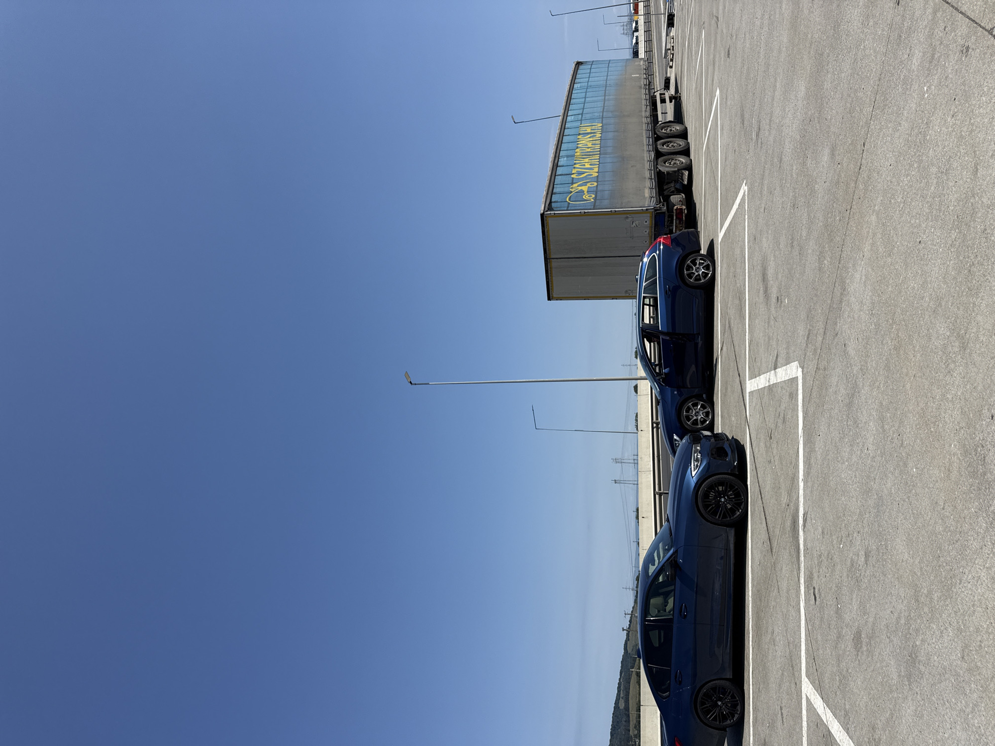 The image depicts an outdoor parking lot on a clear, sunny day. In the foreground, two dark blue cars are parked in designated spots marked by white lines. The car on the left is a sedan, and the car on the right is a station wagon with its driver's side door open. Behind the cars is a large, gray semi-trailer truck with yellow lettering that reads "SZAKITRANS.HU". The truck has multiple axles and appears to be stationary. In the background, a guardrail separates the parking lot from what appears to be a highway or elevated road. Beyond the guardrail, rolling hills covered in vegetation are visible under a bright blue sky. Several tall, metal light poles are also present throughout the scene. The location is identified as Sanpetru, Romania.