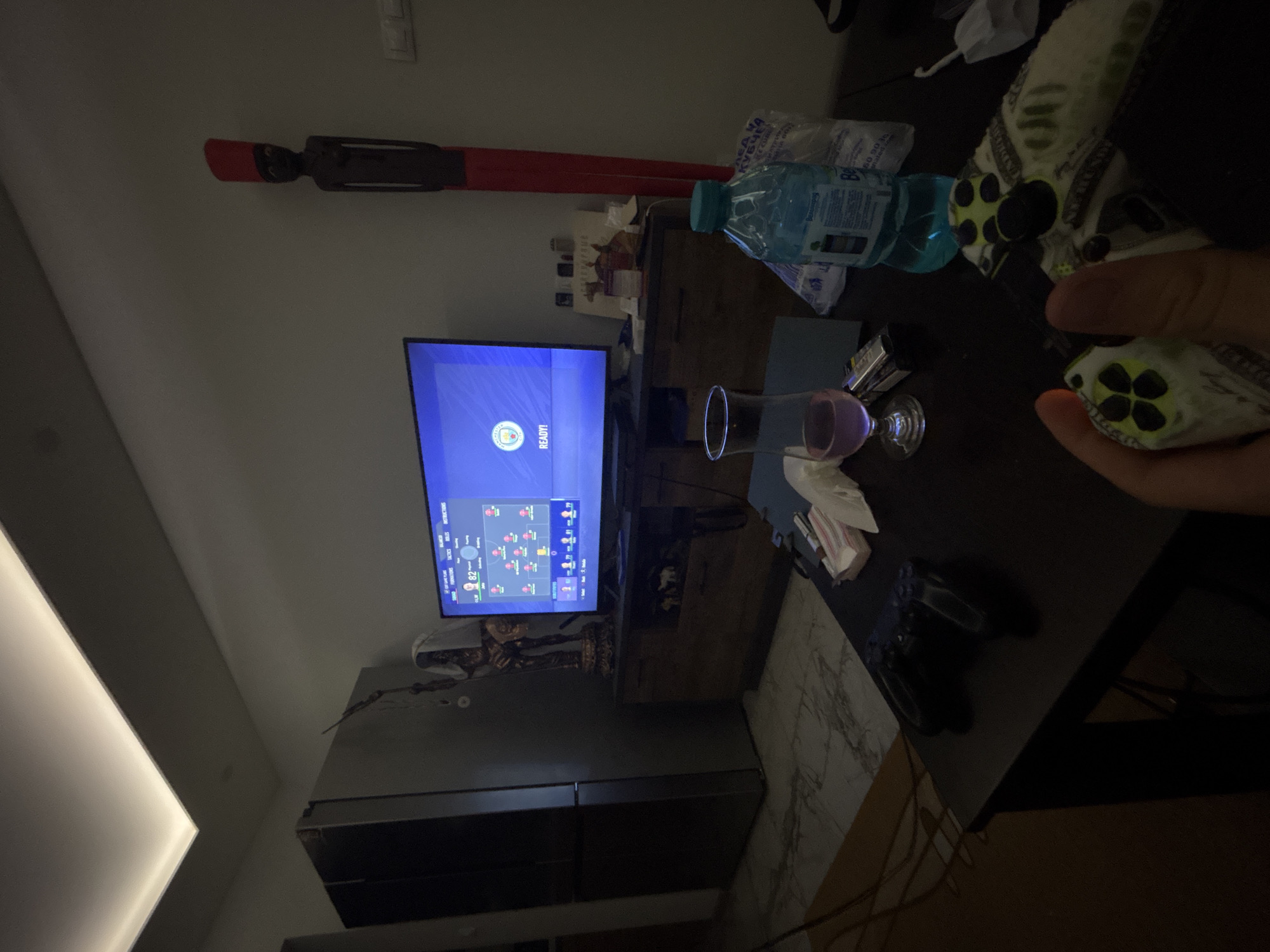 The photograph shows an indoor scene at night. A television displays a game screen with the word "READY!" visible. There is a glass of liquid on a table, along with a bottle of water, tissues, and a game controller. A person's hand is holding a controller. A statue is visible below the TV. The location is in Sofia, Bulgaria.