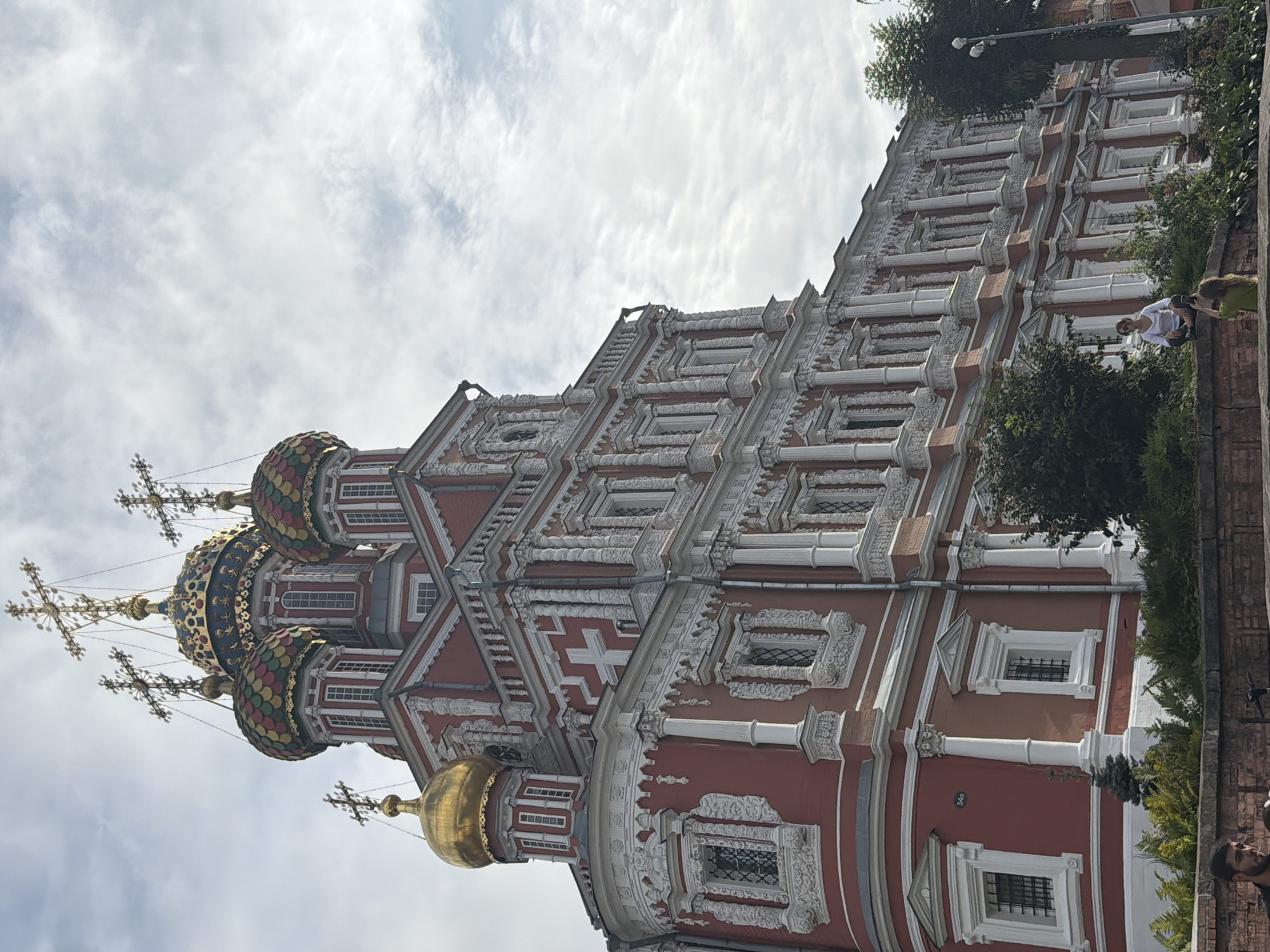 The image captures an exterior view of the Cathedral of Saints Peter and Paul in Nizhny Novgorod, Russia, on a cloudy day. The cathedral is a striking red building adorned with white ornate decorations and multiple domes. Several of the domes are gilded or covered in colorful tiles, topped with crosses. In the foreground, two people are visible. One, a woman in a white top, sits on a low brick wall, looking towards a second person. The second person, also a woman, is partially visible, facing away from the camera and appearing to hold a phone to take a picture. The area around them is landscaped with greenery.