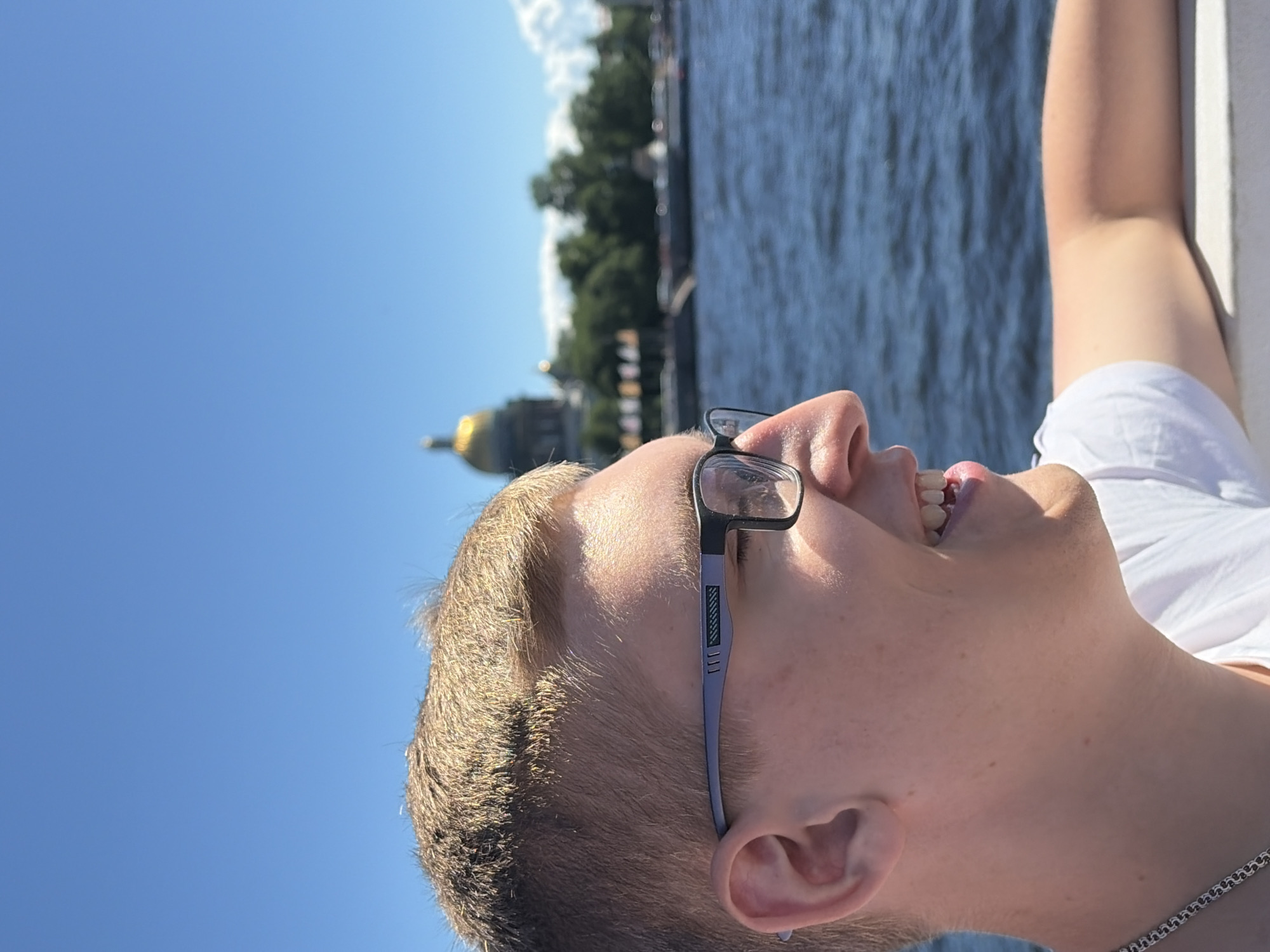 The image shows a close-up profile view of a young white male with blond hair and glasses, smiling. He is wearing a white t-shirt and a silver chain necklace. The background is a bright blue sky and a body of water, likely a river or canal. In the distance, a golden-domed building, possibly St. Isaac's Cathedral, is visible, along with trees and other buildings. The overall scene suggests a sunny day in a city with notable architecture. The person appears to be on a boat or near the water. The scene is set in Russia, likely St. Petersburg, given the architectural landmark.