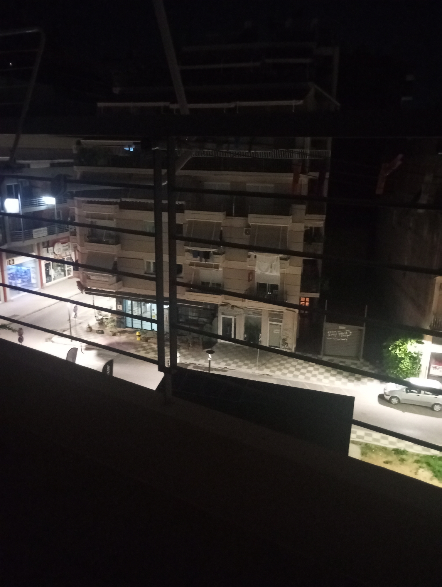 This image captures a nighttime street scene viewed from a slightly elevated perspective, likely a balcony or window, through a series of horizontal bars. In the foreground, dark, silhouetted structures partially obscure the view. Beyond these bars, a multi-story building with a light-colored facade dominates the mid-ground. The building has multiple windows, some illuminated, revealing interior spaces. Balconies with railings are visible on several floors.
Below the building, a street runs horizontally across the scene. The street is paved with checkered tiles in some areas and asphalt in others. Several lampposts are present, casting pools of light onto the pavement. A white car is parked on the right side of the street. On the left side of the street, the facades of shops or businesses are visible, with some illuminated windows displaying products. A small patch of green vegetation is present on the right side of the street near the car. The overall impression is that of an urban street at night, with the presence of buildings, a street, and parked vehicles. The scene is located in the Municipal Unit of Agrinio, Greece.