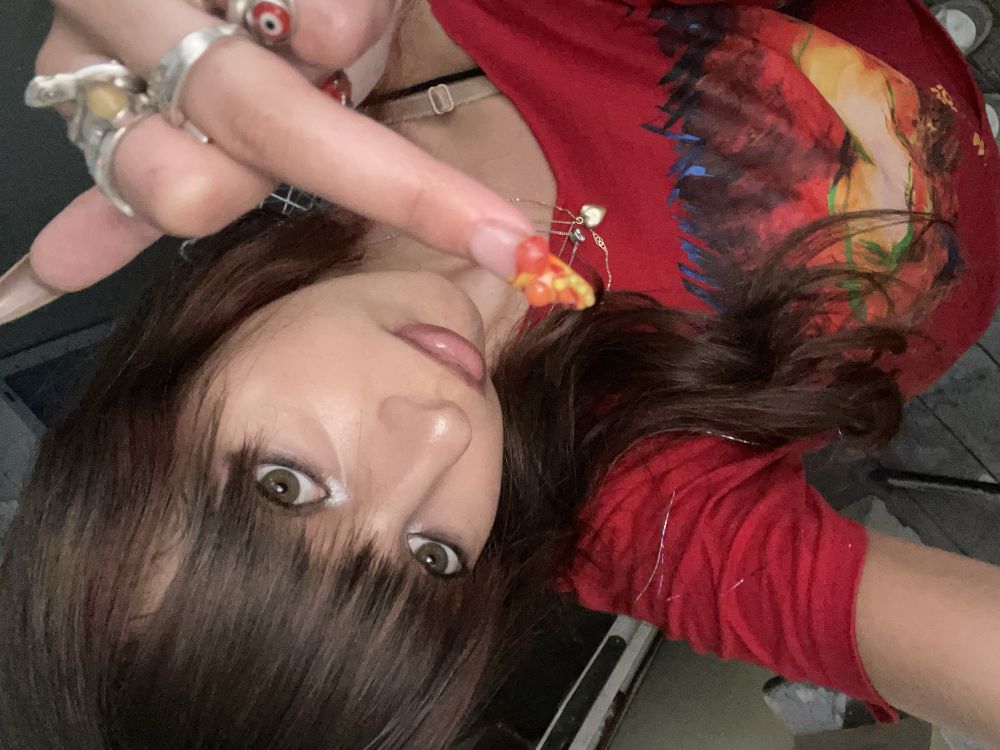 A close-up, medium shot shows a young woman with brown hair and bangs, wearing a red t-shirt with a colorful abstract design. She is looking directly at the camera, her left eye slightly closed. Her right hand is raised, with long, painted fingernails and several rings on her fingers, including one with an evil eye design. Her left hand is visible on her chest, with a small heart-shaped pendant on a necklace. The background is dark and out of focus, suggesting an indoor environment. There is no discernible information about the location in Raanana, Israel.