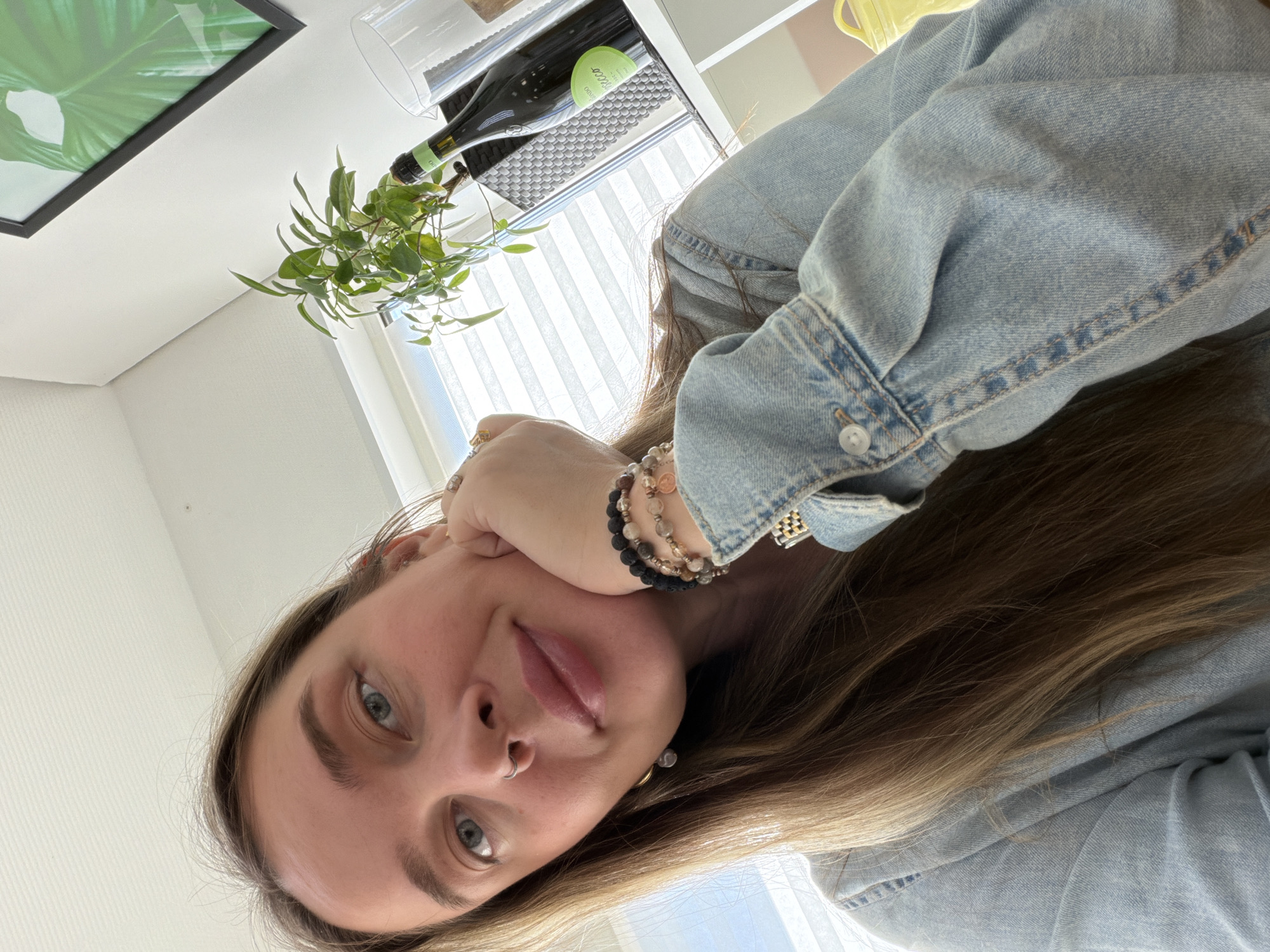 A selfie shows a young woman indoors. She has fair skin, light brown hair, and blue eyes. She is wearing a light blue denim jacket and several bracelets on her left wrist. She is resting her left hand on her face. A bottle of wine, a clear glass, and a small green plant are visible in the background. A framed print of a green plant is partially visible in the upper left corner. Venetian blinds are visible through a window in the background. The location is not specified in the image and cannot be identified.