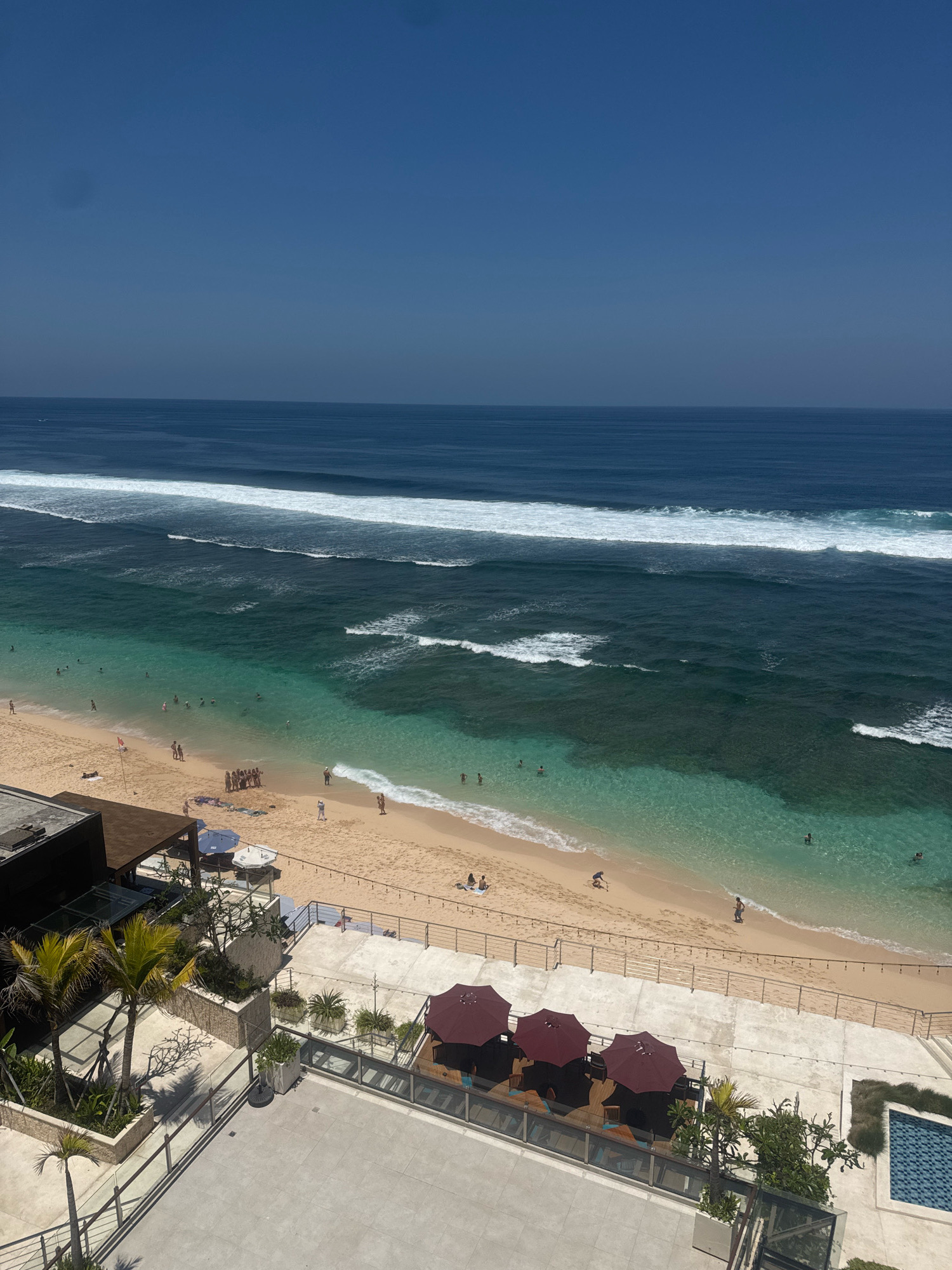 The image captures a high-angle view of a tropical beach resort area in Kuta Selatan, Indonesia. The scene is divided into distinct zones: the ocean, the beach, and the resort amenities.
The ocean dominates the upper half of the image, transitioning from a deep blue to lighter turquoise hues closer to the shore. White-capped waves create a prominent line of surf parallel to the coastline. Scattered individuals are visible in the water, some appearing to swim or wade.
The beach is a wide expanse of light-colored sand, populated with several small groups of people engaged in various activities. Some are walking along the shoreline, others appear to be sunbathing or socializing. A red flag is planted in the sand near a cluster of people.
Adjacent to the beach is a paved resort area with a row of dark red umbrellas providing shade over seating areas. Several palm trees and potted plants are interspersed throughout this space, adding to the tropical ambiance. In the lower right corner, a small tiled swimming pool is visible. The overall impression is a sunny, tranquil beach resort scene.