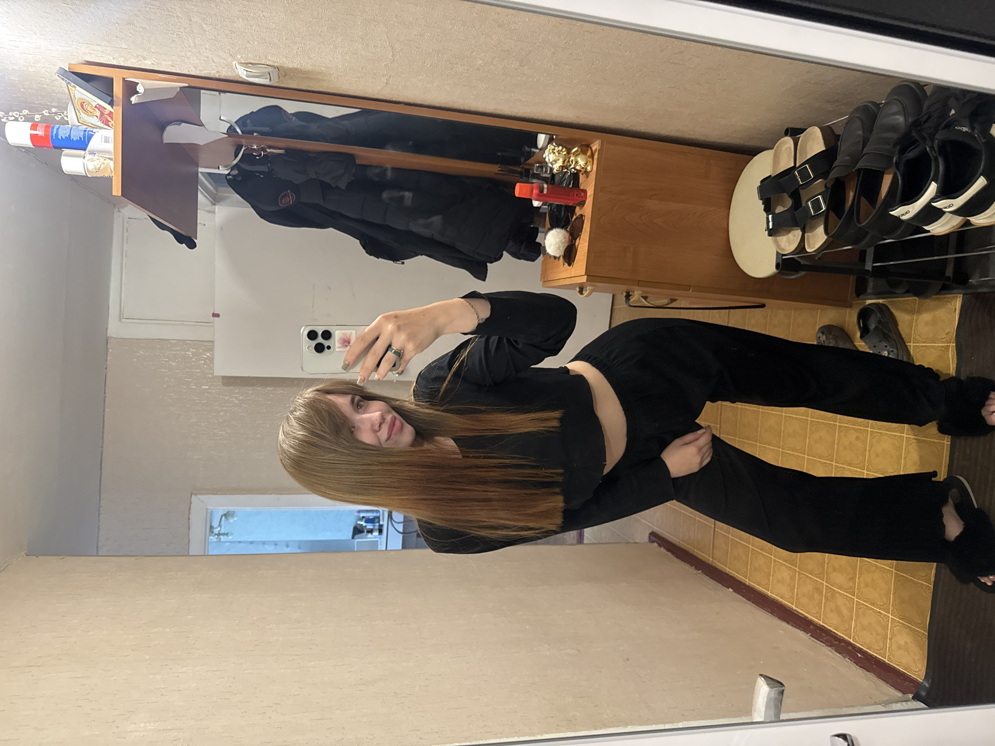 A woman is taking a mirror selfie in what appears to be an entryway or hallway. She is wearing a black long-sleeved crop top and black pants. The mirror reflects a coat rack with clothes hanging on it, a shelf with various items, and a shoe rack with shoes. The floor has a tile pattern. The window shows some sort of building that would potentially allow location to be the city of Chaikovsky, Russia.