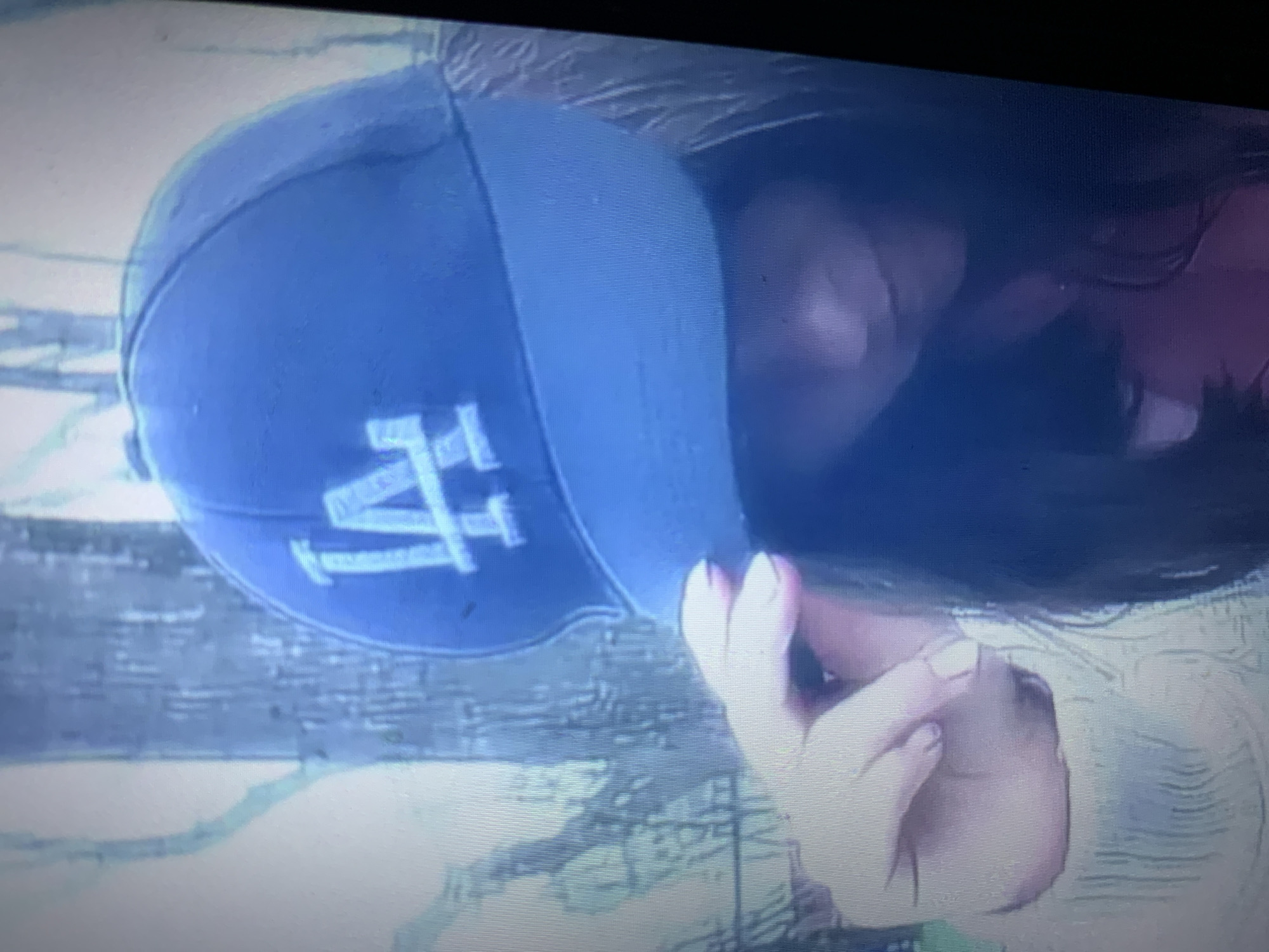 A low-angle, medium shot shows a person wearing a dark blue baseball cap with a light blue brim. The cap has a white "LA" logo. The person is holding the brim of the cap with their right hand, and their face is mostly obscured by the cap and their dark hair. They are wearing a light-colored, textured shirt. The background is out of focus and appears to be outdoors, with hints of trees or structures. The image is grainy and has a blueish tint, suggesting it may be a photograph taken from a screen. There are no specific details in the image that indicate a location in the city of Lyman, Ukraine.