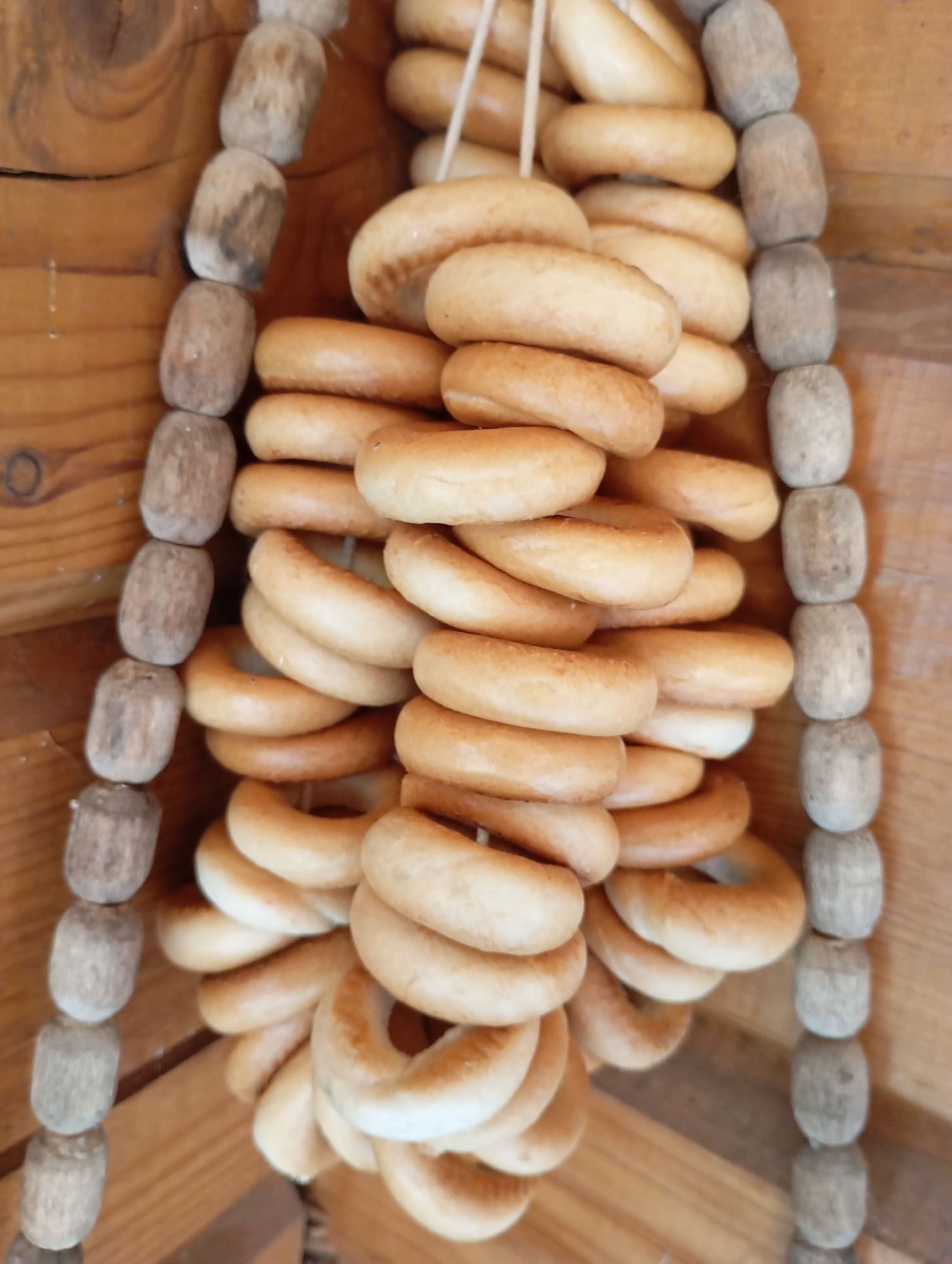 The image shows a close-up, vertically oriented view of several strings of baked goods, likely bagels or a similar ring-shaped bread product, hanging against a wooden background. The baked goods are light brown and appear to be threaded onto thin white strings. Interspersed between the baked goods on some of the strings are cylindrical, wooden beads, also strung together. The wooden background consists of planks of varying widths and colors, suggesting a rustic setting, possibly an exterior wall or an interior wooden structure. The lighting is relatively even, highlighting the textures of both the baked goods and the wood. There are no people or animals visible in the image. The scene appears to be static, with the baked goods hanging still. The location is identified as Malyi Bukor, Russia.