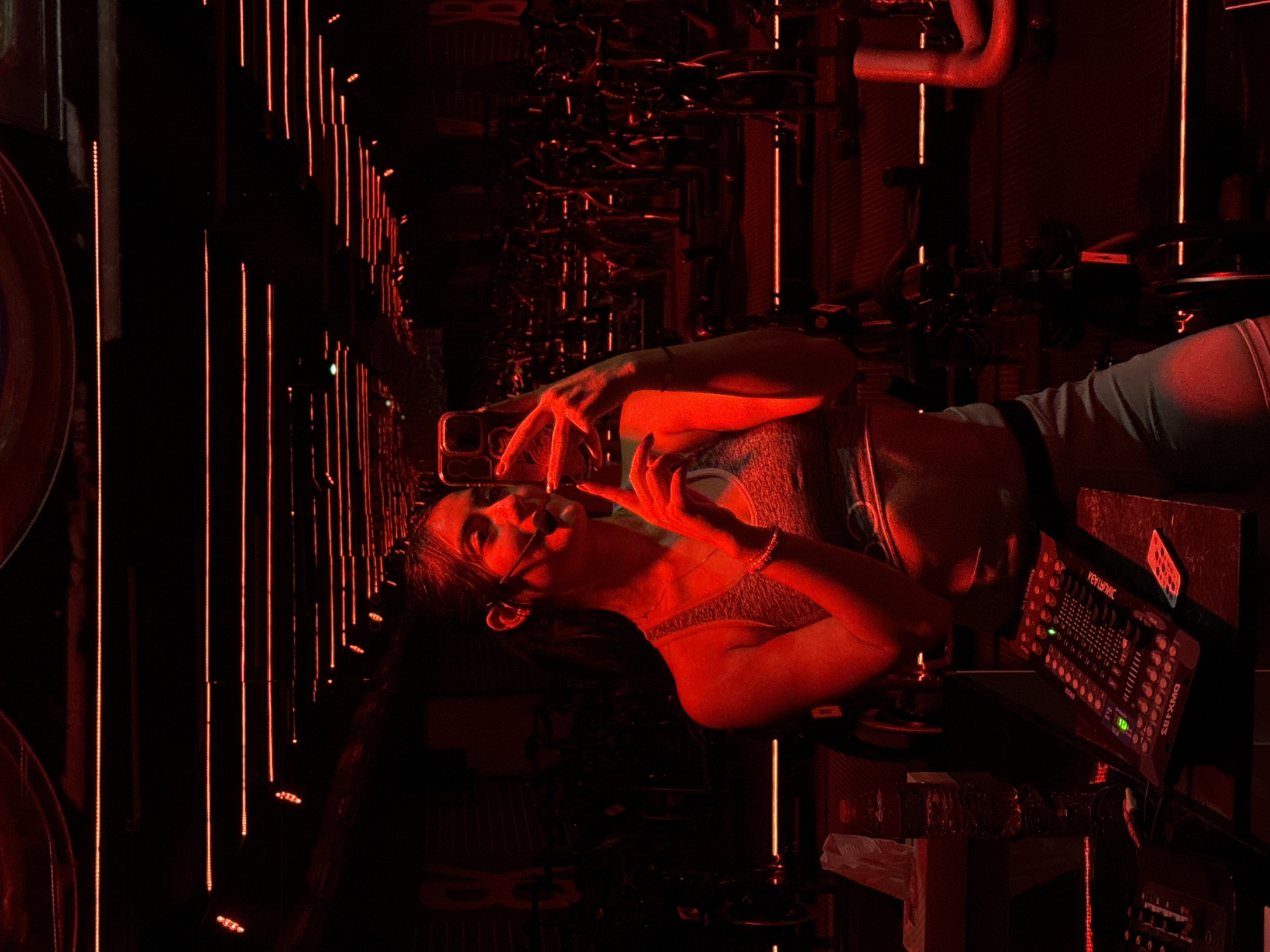 A dimly lit, red-toned interior with rows of spin bikes is captured, likely a spin studio, or gym in Puebla, Mexico. A woman is positioned in the foreground, taking a photo of herself with her phone. She's wearing a sports bra and workout clothes. In the lower right of the frame is a mixing board, control unit, a bottle of water, and additional audio equipment. Red vertical light strips illuminate the space between the rows of bikes.