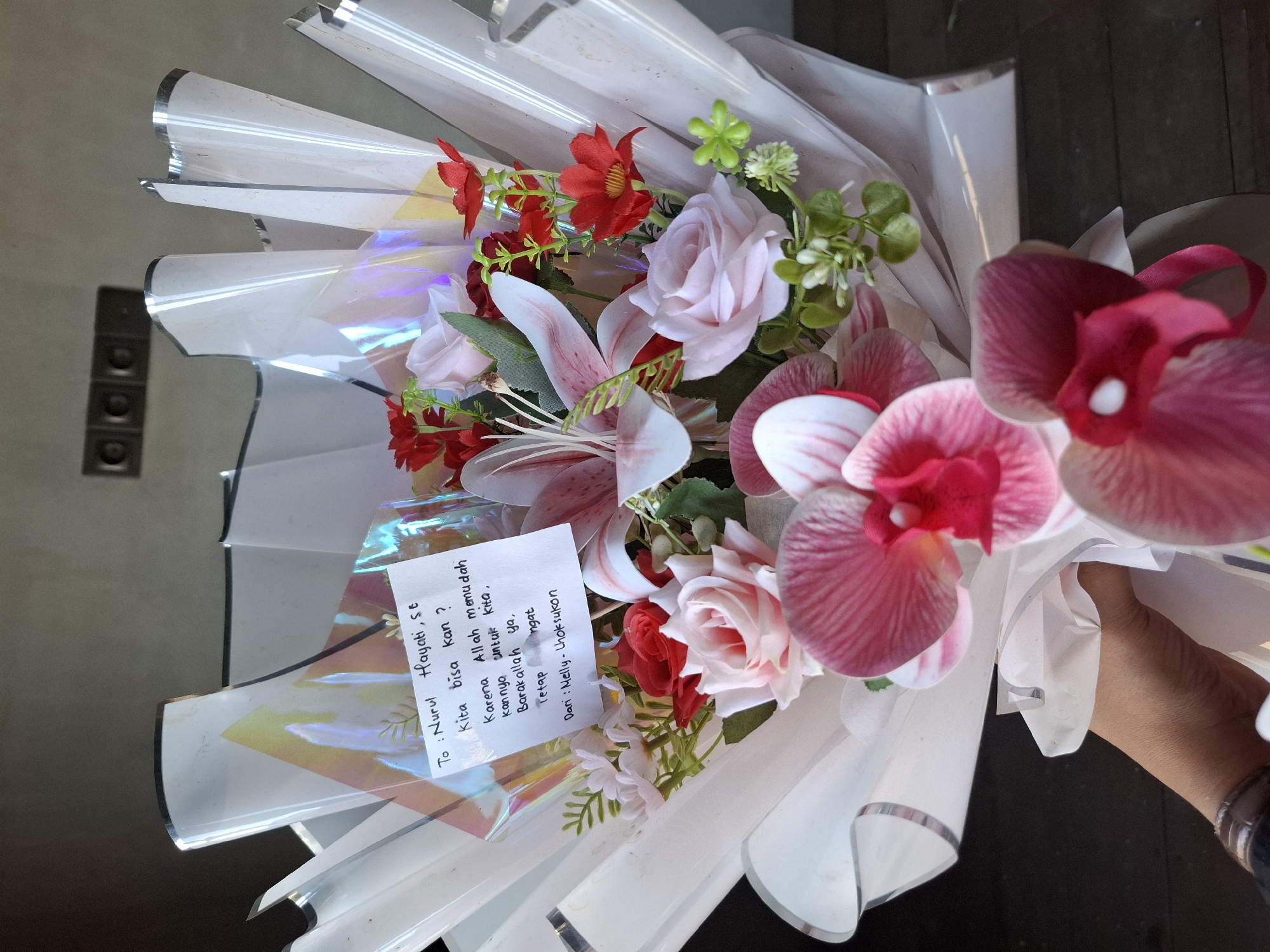 The image shows a bouquet of artificial flowers, primarily pink and red, wrapped in clear plastic with a iridescent sheen. A handwritten note is attached to the bouquet, written in Indonesian. The note reads: "To: Nurul Hayati, S.E. Kita bisa kan? Karena Allah memudahkan jalannya untuk kita, Barakallah ya. Tetap semangat. Dari: Melly - Uhtsukon". The bouquet is being held by a hand, partially visible at the bottom left of the frame, wearing a watch with a brown strap. In the background, a wall with two electrical outlets is visible, suggesting an indoor setting. The lighting is dim, creating shadows. There is no indication of the specific location being Gronggrong, Indonesia.