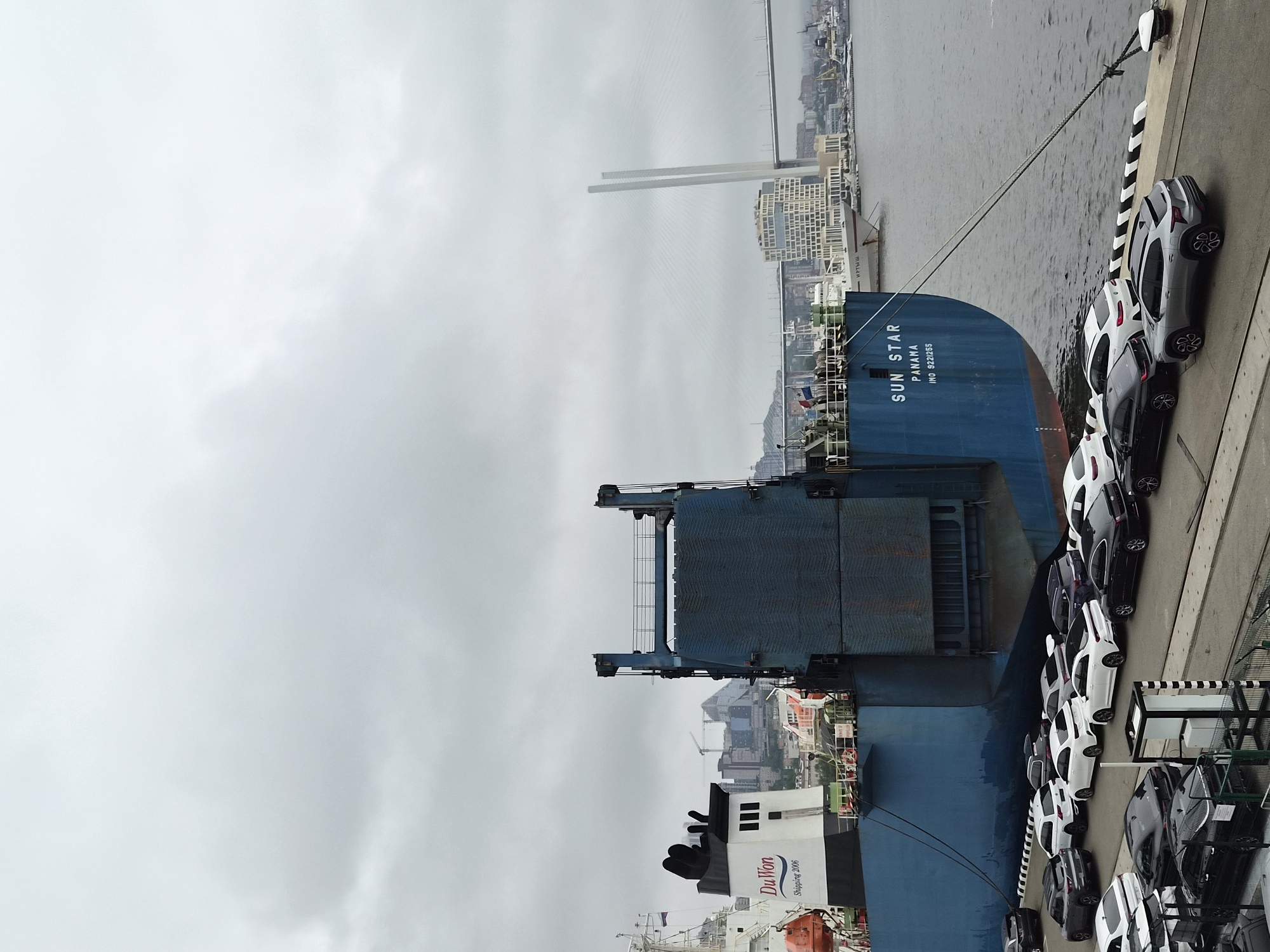 This is a medium-distance, eye-level, overcast shot of a large blue car carrier ship named "SUN STAR" docked at a port in Vladivostok, Russia. The ship is emblazoned with "SUN STAR PANAMA INO 9221255" in white lettering. Cars are parked on the pier in front of and beside the ship, with most appearing to be new. The pier is lined with black and white striped bollards. In the background, the Zolotoy Rog Bay is visible, with bridges and city buildings on the horizon. The sky is cloudy and gray. There are no people visible in the frame.