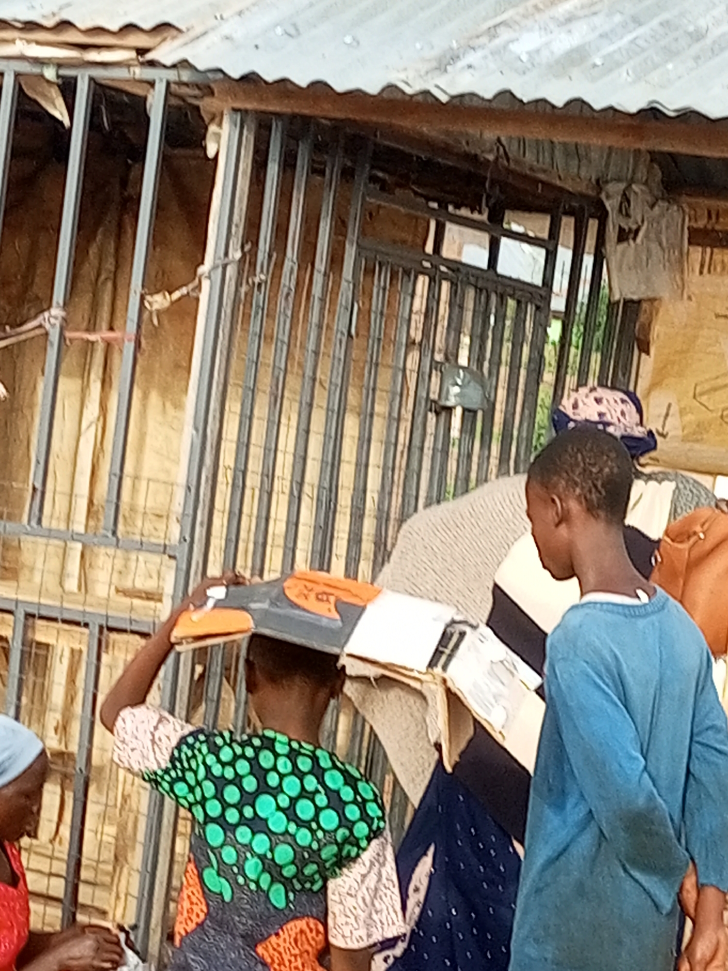 In Tumfure, Nigeria, a young person in a blue shirt stands with their hands behind their back, facing away from the camera. To their left, another young person in a green and black patterned top carries a tray on their head. A third person, wearing a red shirt and a blue head covering, is partially visible to the far left, engaged in an activity with their hands. The background consists of a corrugated metal roof over a structure with metal bars and tan-colored sheeting, suggesting an outdoor market or stall. The overall scene depicts daily life and commerce in a Nigerian community.