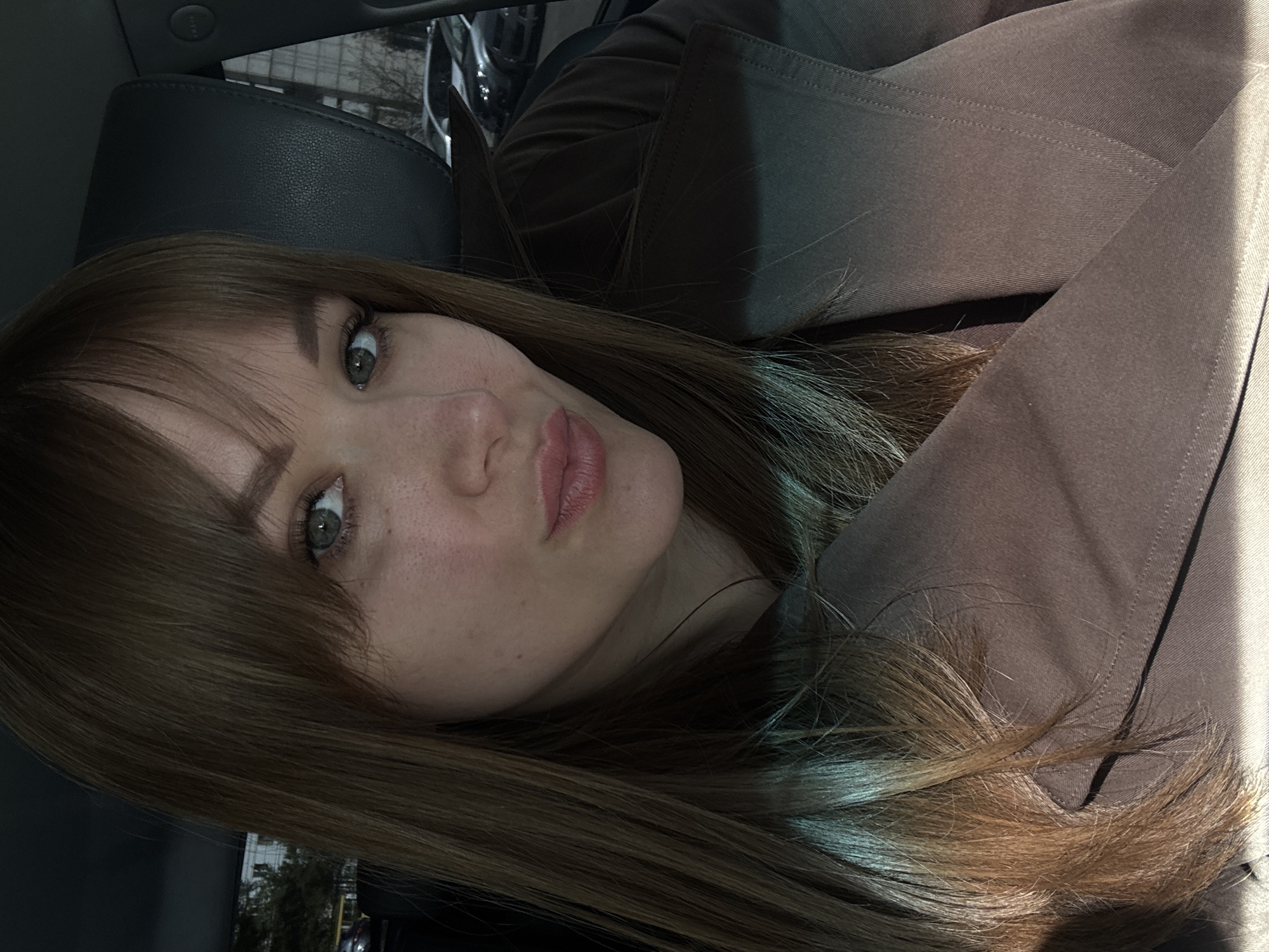 A woman with long brown hair and bangs is depicted in a close-up, slightly angled shot. She has light skin, grey eyes, and pink lips. She is wearing a brown trench coat and appears to be in a car, based on the black leather car seat visible in the background. The background is blurry, suggesting movement or out-of-focus elements. The picture includes a glimpse of a city scene through the car window, possibly indicating an urban setting. Without additional information, determining the exact location within Chaikovsky, Russia, is impossible.