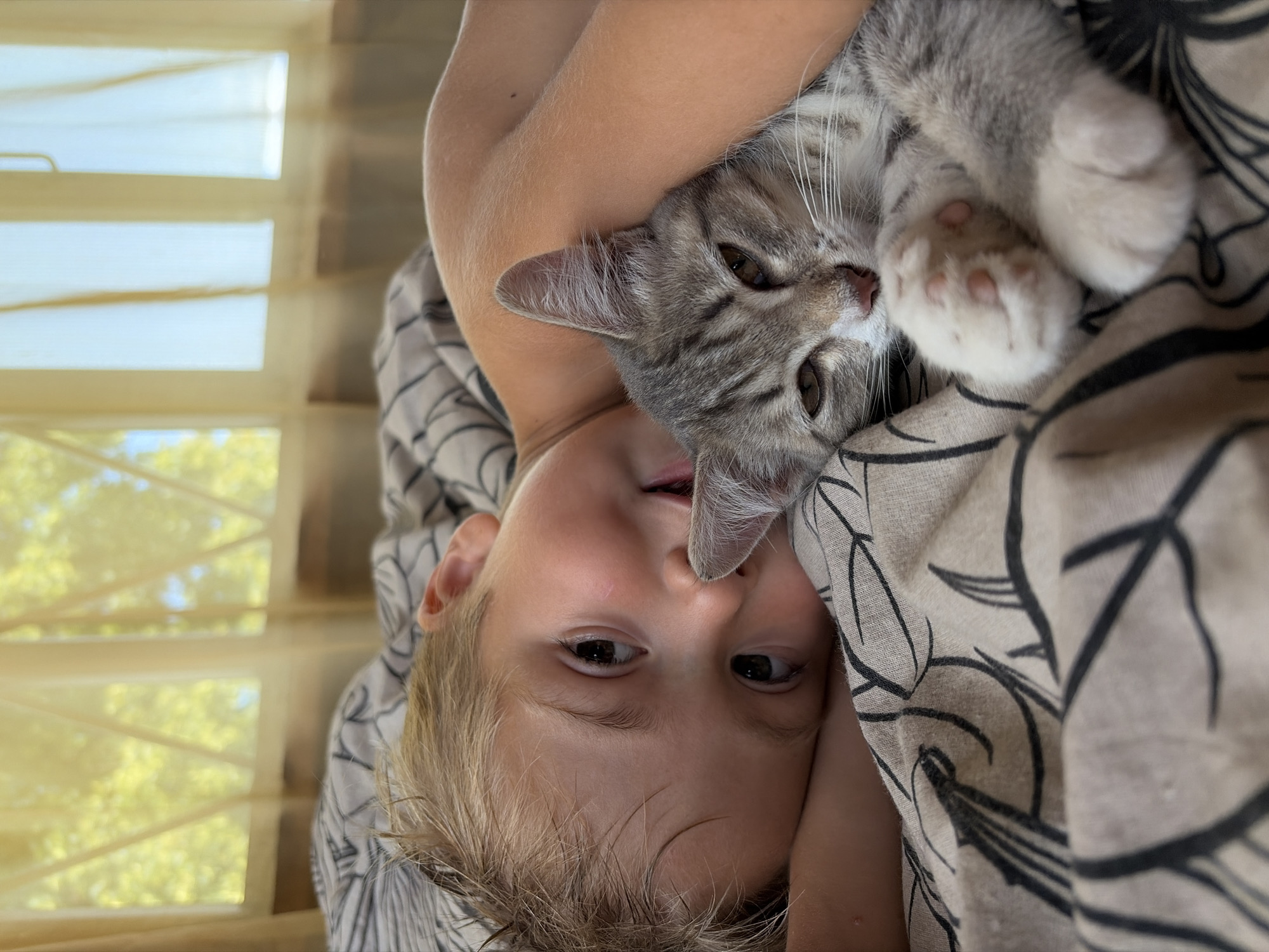 A close-up, high-angle shot captures a young boy with blonde hair lying in bed, his head propped up by a pillow. He is looking towards the camera with a slight smile, his right arm around a tabby cat that is resting beside him. The cat is curled up, facing away from the camera, with its paws tucked beneath its chin. The scene is set indoors, with sheer curtains visible in the background, suggesting a window that lets in natural light. The bedding is patterned with black and white stripes and floral designs. The boy's skin appears fair, and the cat has a grey and white striped coat. The overall mood is one of comfort and affection between the boy and his pet. The image is set in a bedroom located in Olkhovka, Russia.