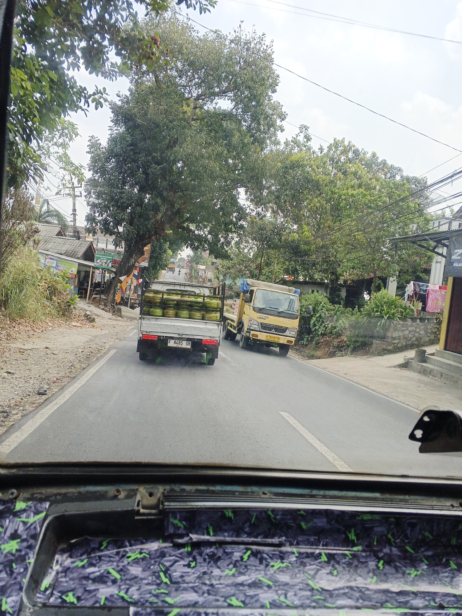 An eye-level, exterior, medium shot shows a slightly hazy, overcast day on a two-lane road in Cikembar, Indonesia. The camera is positioned inside a vehicle, looking forward through the windshield and over the dashboard. A white Daihatsu truck, laden with bright green gas canisters, is in the foreground, traveling in the same direction as the viewer. To its right, a yellow Mitsubishi Fuso truck is also in motion. Both trucks have Indonesian license plates.

The road is paved and has a white dividing line. On the left side of the road, there is a gravel shoulder, dry grass, and a row of small shops with peaked roofs. A large tree with dense foliage arches over the road in the middle distance, partially obscuring the sky. On the right side of the road, there are more trees, houses, and laundry hanging to dry. Power lines crisscross the sky. The foreground of the image is dominated by the interior of the viewing vehicle, showing the dashboard and windshield frame, with a patterned surface visible on the dashboard. The overall impression is of a busy, rural road scene with commercial activity.