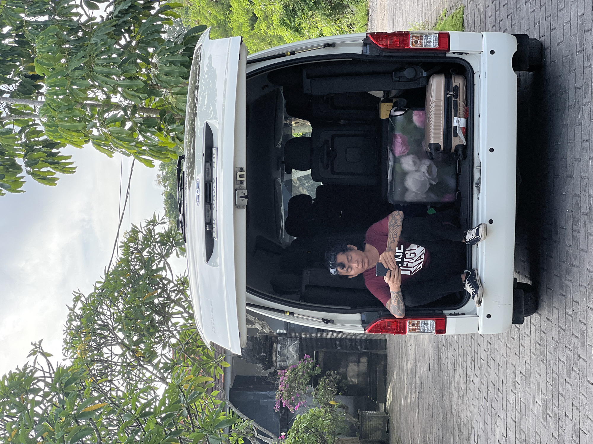 An adult male is relaxing inside the open tailgate of a white van or large SUV, parked on a paved driveway in Kuta Selatan, Indonesia. He is sitting with his legs extended onto the vehicle's bumper, casually looking at a dark-colored smartphone held in his hands. He wears a dark red short-sleeved t-shirt with a graphic design, dark pants, and dark sneakers. Tattoos are visible on both of his arms, and a pair of sunglasses is pushed up onto his dark hair.
Inside the vehicle's spacious cargo area, two pieces of luggage are visible: a small, light brown hard-shell suitcase and a larger, soft-sided suitcase with a floral or abstract pink and white pattern. The tailgate is propped open. Faint text "HIACE" can be seen on the side of the vehicle near the tailgate.
The scene is set outdoors during what appears to be daytime, under an overcast sky. Lush green trees and foliage are abundant in the background, with some pink and purple bougainvillea flowers adding color near a traditional-style building with dark, ornate details. The ground is covered with light-colored brick paving. The man appears calm and at ease, perhaps taking a break during travel or an outing. On his t-shirt, the partially visible text reads "THE UGLY MAN."