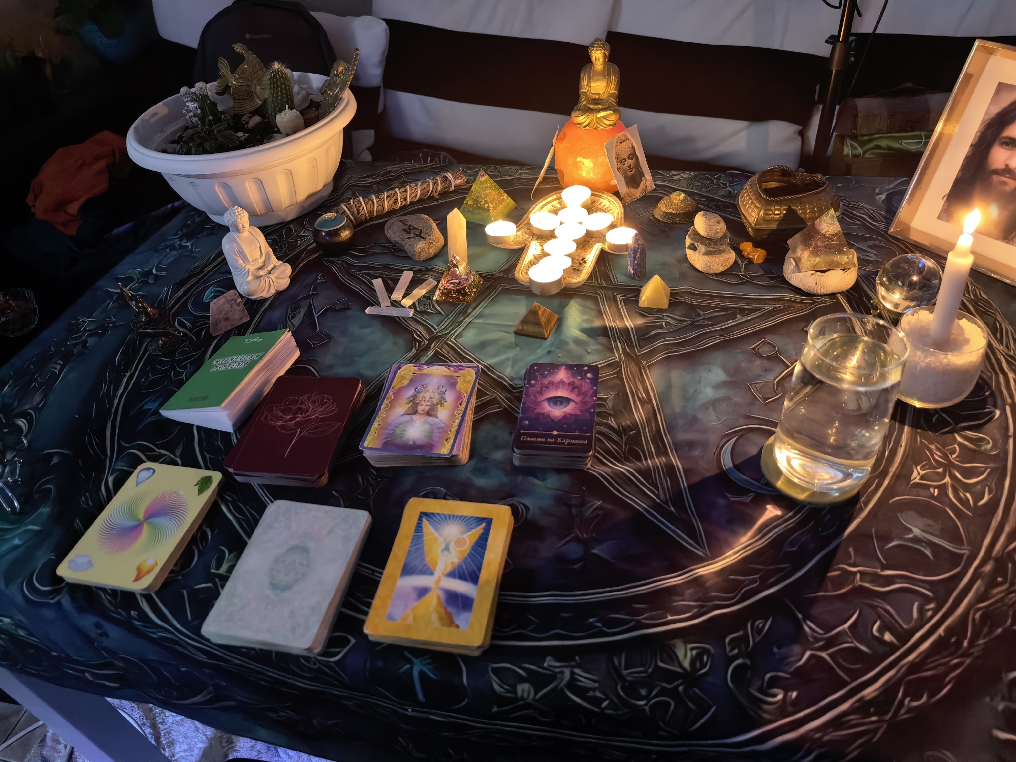 An overhead, slightly angled shot reveals a spiritual or mystical altar set up on a dark, intricately patterned tablecloth. The scene is dimly lit, primarily by numerous candles, suggesting evening or nighttime.
In the background, a white planter filled with succulents, cacti, and small decorative golden fish is visible. To its right, a white Buddha statue sits serenely. Further right, a golden Buddha statue rests atop an illuminated orange object, likely a salt lamp. A framed picture of Jesus Christ stands to the far right.
The central area of the table is adorned with various spiritual items. A bundle of sage rests near several crystals, including clear points, pyramids (one green, one brown), and tumbled stones. A tray holds multiple lit tea light candles, casting a warm glow. Another tall, white taper candle burns brightly to the right. A glass of water sits nearby.
In the foreground, several decks of oracle or tarot cards are neatly stacked. Visible text on a green book reads "КУРИНЫЙ БУЛЬОН" (Chicken Soup). One purple card deck has "Пътят на Кармата" (The Path of Karma) printed on it, and another partially visible deck appears to have "АНГЕЛИТЕ" (The Angels) on its packaging. The overall atmosphere is serene and contemplative, indicative of a space used for meditation, divination, or spiritual practice.