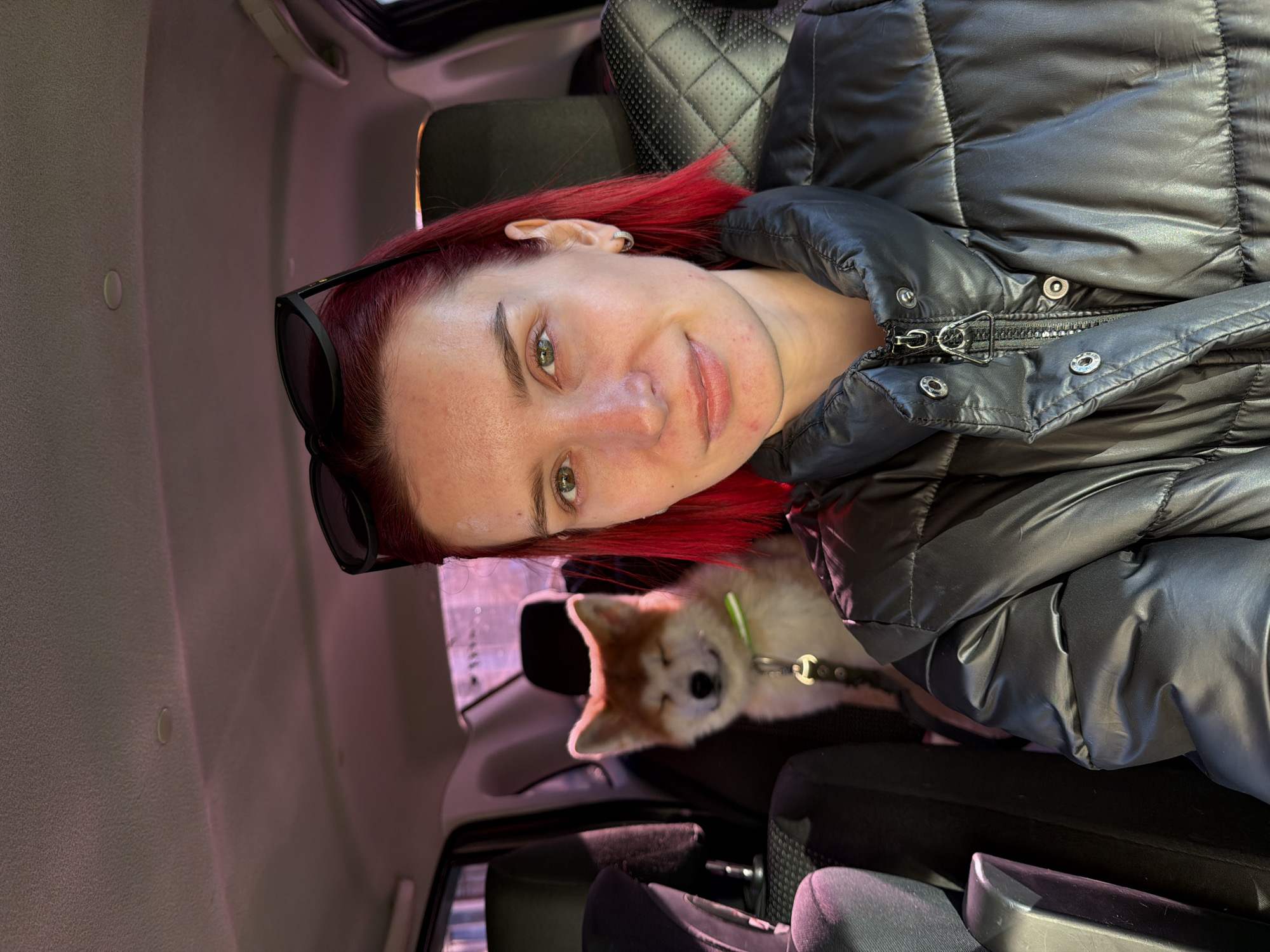 A young woman with striking red hair and green eyes is captured in a close-up photo inside a vehicle. She has fair skin with some minor blemishes and a slight, friendly smile, looking directly at the camera. Black sunglasses are perched on her head, and she wears a silver hoop earring. Her attire includes a black, glossy puffer jacket with visible zippers and snap fasteners.
Beside her, a light-colored dog, possibly an Akita puppy, is visible, wearing a lime green collar and a black leash. The dog appears calm, resting or sitting quietly.
The setting is the interior of a car or SUV, indicated by the black quilted seats and a light-colored headliner. Light from outside, tinged with a soft purple-pink hue, filters into the vehicle, suggesting it is daytime, perhaps late afternoon or an overcast day. The overall scene conveys a casual, relaxed moment shared between the woman and her pet while traveling or resting in the car. There is no visible text. The location context for the image is Chaikovsky, Russia.