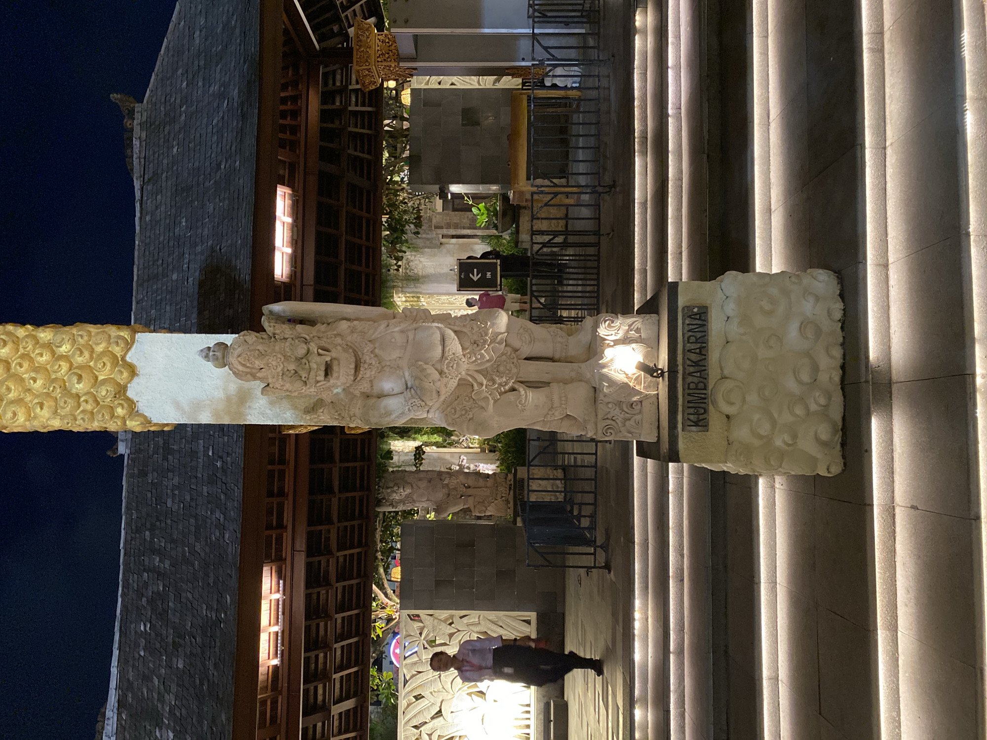 A statue of Kumbhakarna, a character from the Ramayana, stands prominently in front of a building at night in Kuta Selatan, Indonesia. The statue is carved from a light-colored stone and is adorned with gold at the top. It is placed on a pedestal with a textured design. The building behind the statue features a dark, tiled roof with visible wooden support beams. Illuminated stairs lead up to the structure, with subtle lighting integrated into the risers.
In the background, two individuals are visible. One, a woman in a light-colored shirt and dark pants, walks from left to right across a paved area. Another person, wearing a red shirt, stands near a sign with an arrow pointing left. A metal fence separates the foreground from the background. The overall scene is dimly lit, with artificial lighting highlighting the statue and the architectural elements of the building.