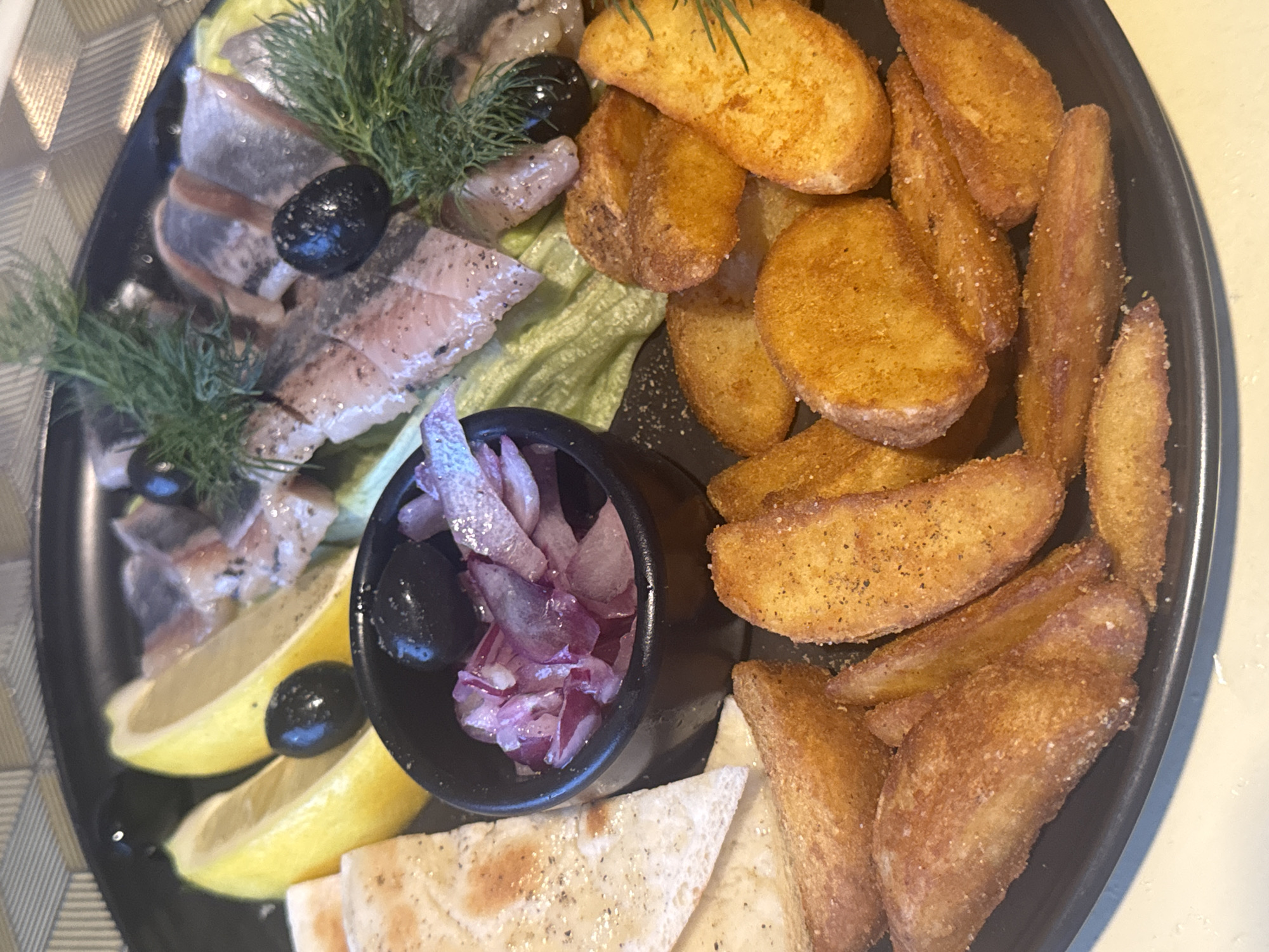 The image shows a platter of food, likely in a restaurant setting. The platter is black and contains several food items. In the foreground, there are numerous potato wedges, seasoned with what appears to be salt and pepper, and some with a reddish spice. To the left of the potato wedges are pieces of flatbread or pita. Behind the flatbread are two lemon wedges and a small black bowl filled with sliced red onions and black olives. The upper portion of the platter features slices of what appears to be pickled herring, garnished with fresh dill. Black olives are scattered amongst the herring. The background shows a textured placemat. The image quality is good, with natural lighting. There are no people visible in the image. This image likely depicts a dish served in Chaikovsky, Russia, given the context provided.