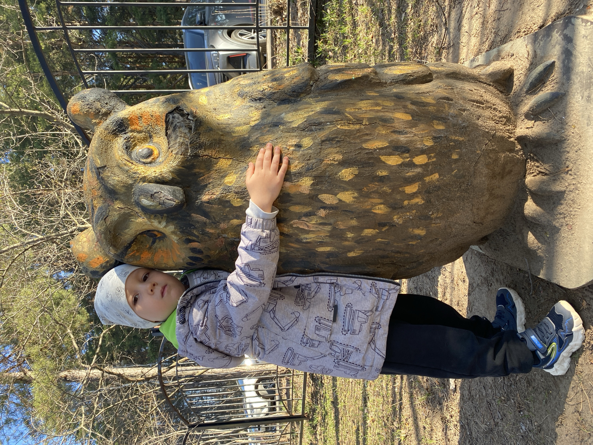 A young child stands next to a large, carved, wooden owl sculpture in Chaikovsky, Russia. The child is wearing a light gray jacket with black construction vehicle designs, black pants, and blue and white sneakers. The child has a white hat and is touching the owl sculpture with their right hand, resting their head against it. The owl is painted with brown, gold, and black details. The background includes trees, a metal fence, and a gray car. The ground is covered with dirt and grass.