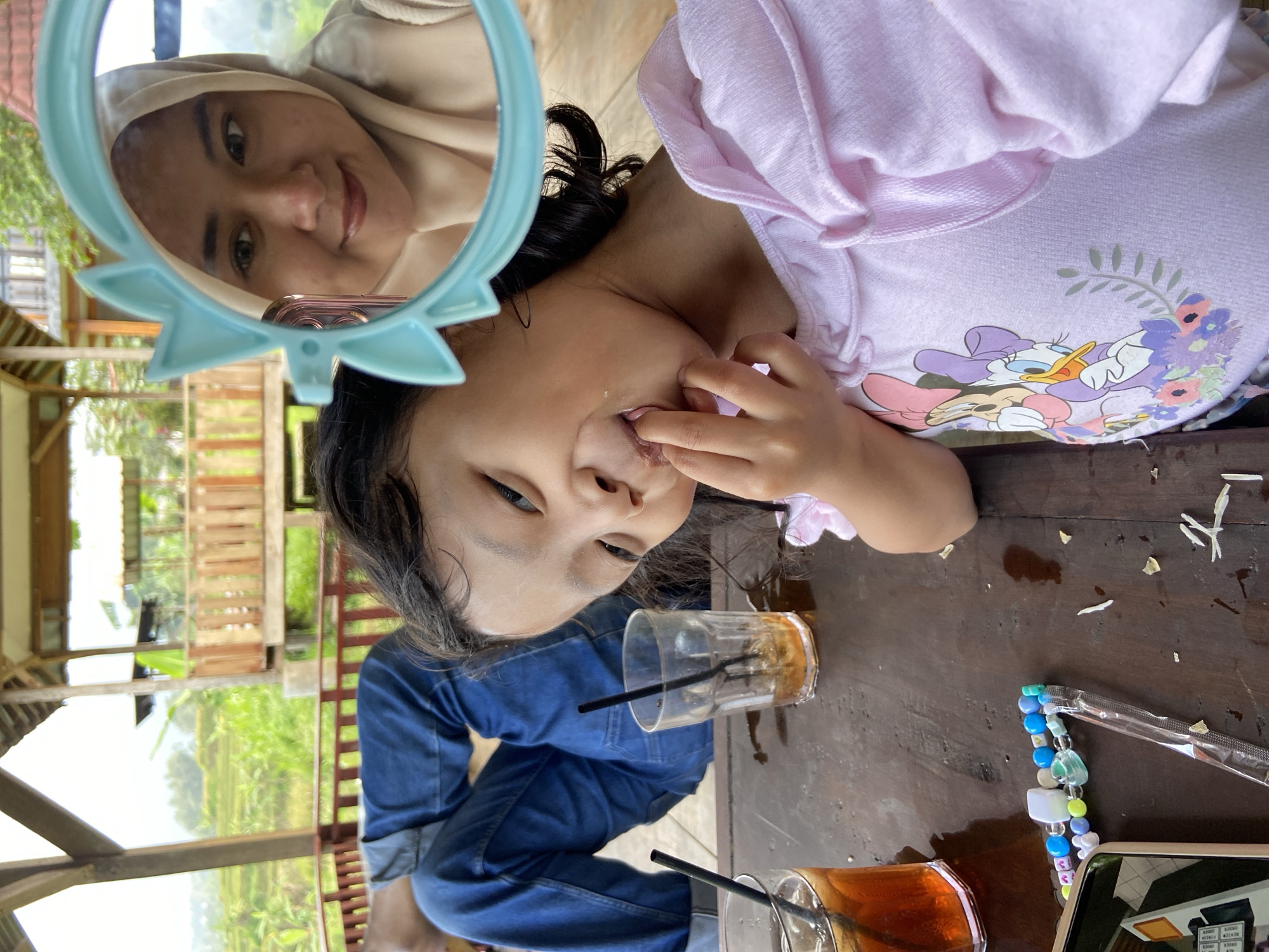 At a table in Tamiajeng, Indonesia, a girl in a pink Daisy Duck sweatshirt sits beside a woman reflected in a blue-framed mirror. The girl touches her mouth. On the table are two glasses of iced tea, a beaded phone strap, and scraps of food. In the background are rice fields seen through the open structure of a building. The girl wears denim.