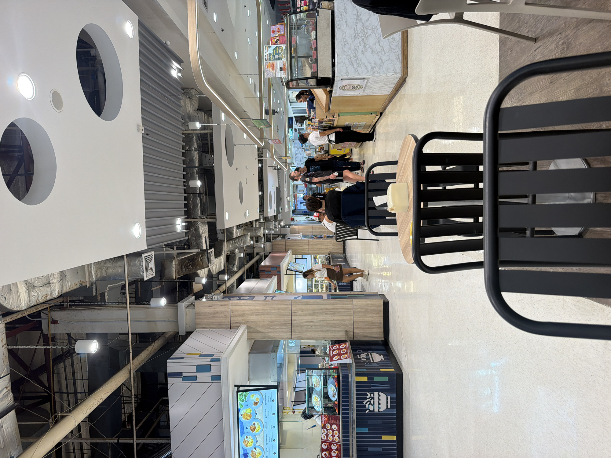 The image shows a food court environment with multiple stalls and seating areas. In the foreground, black metal chairs and a round table with a yellow napkin dispenser are visible. Further back, a food stall with a blue and white striped facade displays menu boards and food items in a display case. A woman in a white top and brown skirt stands near this stall. Beyond this, another food stall features a counter with food items in a glass case and a woman in a white uniform serving customers. People are seen standing and walking around the food court, with various tables and chairs scattered throughout the space. The ceiling is a mix of exposed pipes, ductwork, and white paneling with circular cutouts and recessed lighting. The floor is a light-colored, reflective material. The overall impression is that of a modern, indoor public dining area. The image is likely taken in a shopping mall or a similar commercial establishment in บ้านบางรักษ์, Thailand, given the presence of various food vendors and a bustling atmosphere typical of such locations.