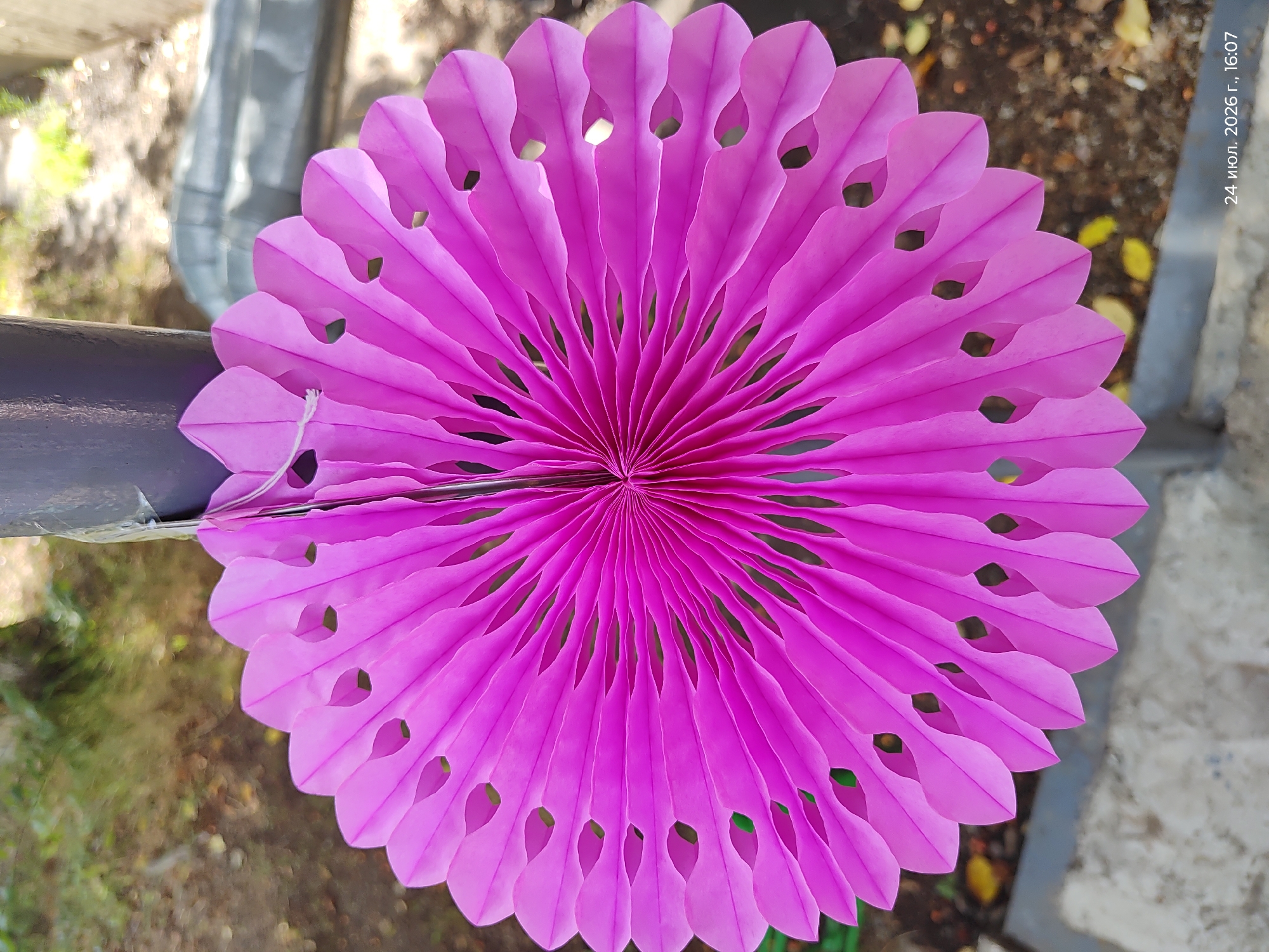 A fuchsia-colored paper fan decoration, featuring cutout petal shapes, is suspended by a white string from a dark gray pole. The fan is fanned out, displaying its pleated texture and radiating from the center. The background is blurred, showing hints of greenery and what appears to be the ground or pavement. A timestamp at the bottom right corner reads "24 июля. 2026 г., 16:07," indicating July 24, 2026, at 4:07 PM.