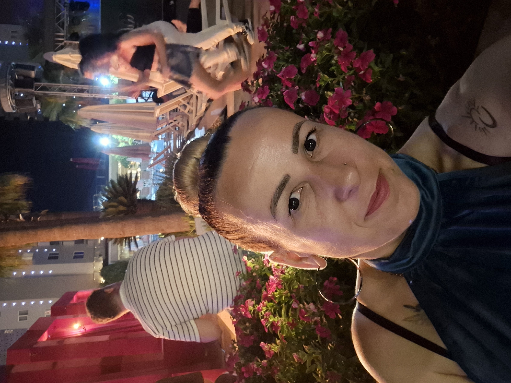 A night-time, eye-level, medium shot shows a woman posing for a selfie in the foreground, with a blurred background of a resort or hotel complex at the beach. The woman, in her mid-30s, has her hair pulled up in a bun and is wearing a dark blue top and large hoop earrings. She has a nose piercing and a tattoo on her left shoulder of a crescent moon and sun. Behind her, a man wearing a striped shirt stands facing away from the camera, and further in the background, two people are dancing. The environment is outdoors, with palm trees, lounge chairs, and a building with lit windows visible.