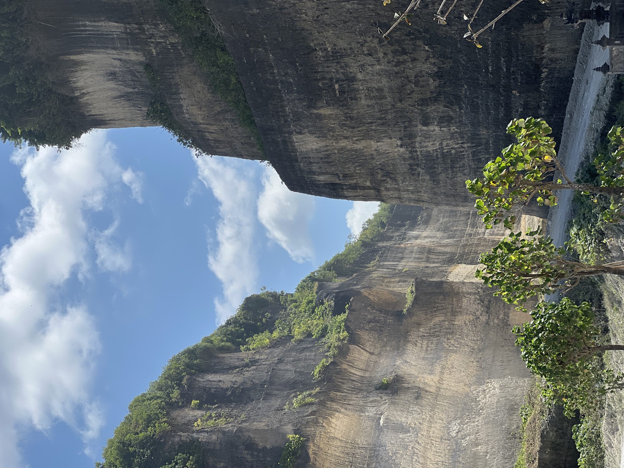 The image displays a majestic gorge or canyon featuring immense, steep rock walls under a bright blue sky with scattered white, fluffy clouds. The rock faces, likely carved from light-colored stone such as limestone, show visible textures, striations, and darker mineral stains. Patches of vibrant green vegetation, including bushes and small trees, cling to the ledges and upper edges of the towering cliffs.
In the lower right foreground, a leafy green tree stands, partially obscuring a stone path or road that runs along the base of the right-hand rock wall. Further along this path, a distinct traditional Balinese temple or shrine with multi-tiered dark roofs is visible, confirming the cultural setting. The massive scale of the rock formations suggests a significant natural or man-made landscape, characteristic of areas like the Garuda Wisnu Kencana Cultural Park in Bali.
The scene is bathed in bright daylight, indicating clear and sunny weather. No individuals or specific activities are discernible, creating a sense of serene grandeur. The architectural style provides a strong contextual cue, aligning with the specified location of Kuta Selatan, Indonesia. No visible text is present in the image.