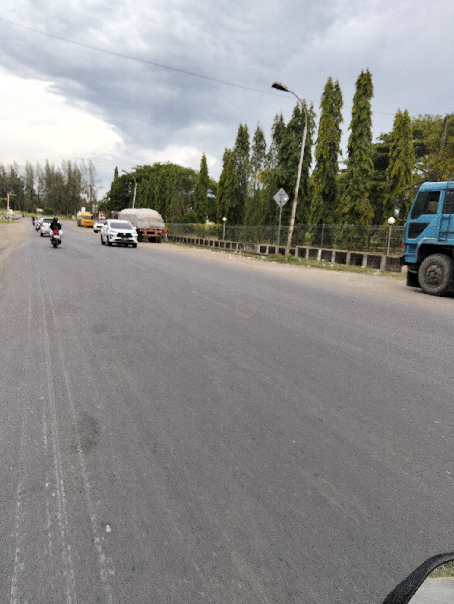 The image depicts a road scene in Tambontunong, Indonesia, with a focus on the road surface and vehicles. In the foreground, the asphalt road shows tire marks and some debris. Several vehicles are visible on the road. To the left, a white car is driving, and further back, multiple motorcycles are in motion. In the background, a large blue truck is parked on the right side of the road. A fence runs along the side of the road, and tall, slender trees line the area beyond the fence. The sky is overcast with gray clouds. The perspective suggests the photo was taken from a moving vehicle.