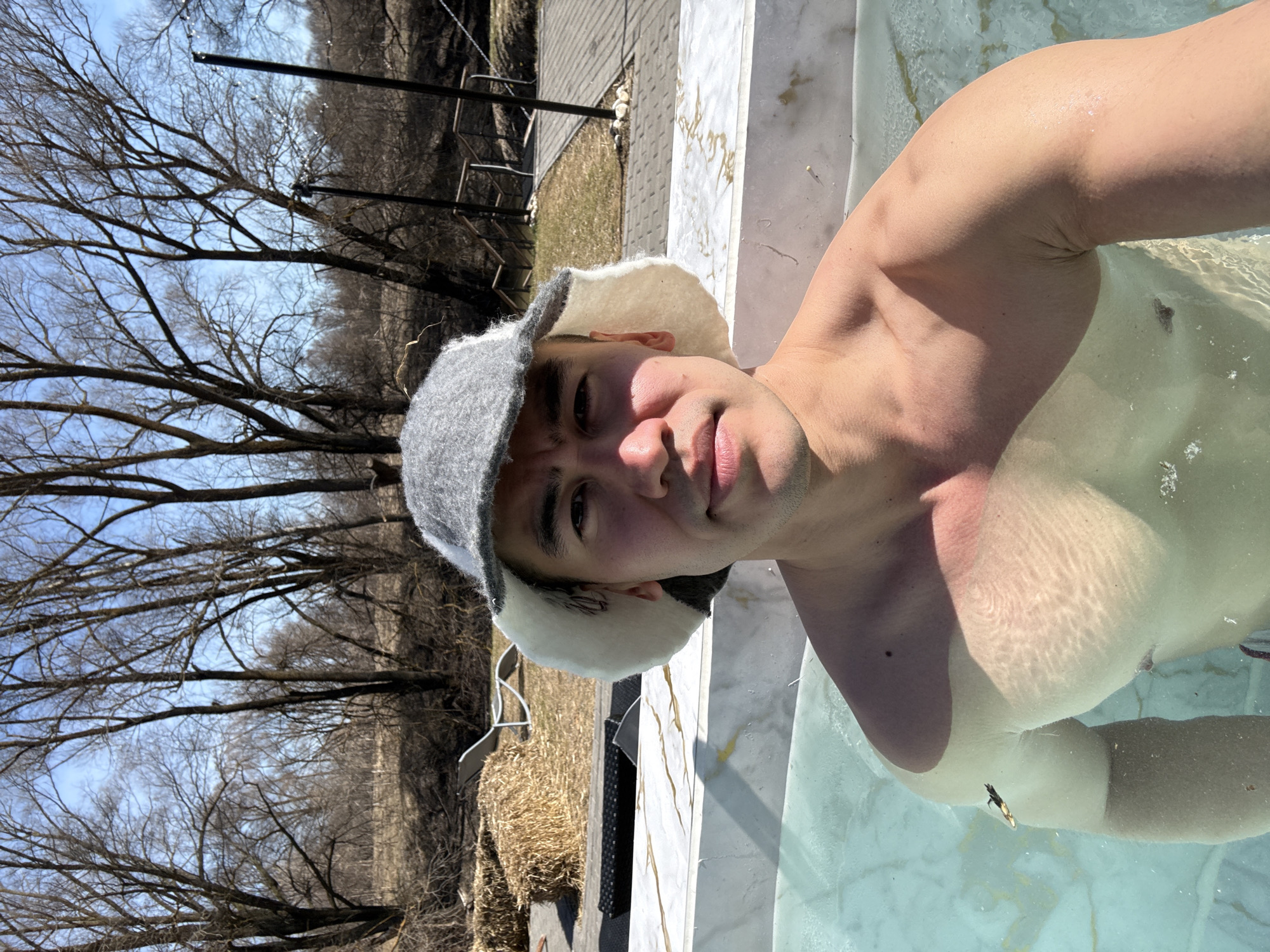 A young man is depicted in an outdoor setting, partially submerged in clear, light blue water. He wears a distinctive grey and white felt hat, commonly associated with Russian banya culture, covering his head and ears. He looks directly at the viewer with a calm, neutral expression. His shoulders, chest, and arms are visible, with reflections of sunlight dancing on the water's surface and his skin.
The man appears to be in a pool or hot tub with a white, marble-patterned edge visible in the foreground. In the background, to his left, are bare tree branches silhouetted against a bright blue sky, suggesting either late autumn, winter, or early spring. Further back, what appears to be a field or grassy area with scattered dry vegetation and some dark structures (possibly a fence or building) can be seen. To the man's right, behind him, a straw bale and a dark outdoor chair are visible on the ground beside the water's edge.
The scene is illuminated by bright daylight, indicating it is likely midday or early afternoon. The clear sky and bare trees suggest a cooler climate, consistent with an outdoor water activity like a banya or hot spring experience in Kazan, Russia. No visible text is present in the image.
