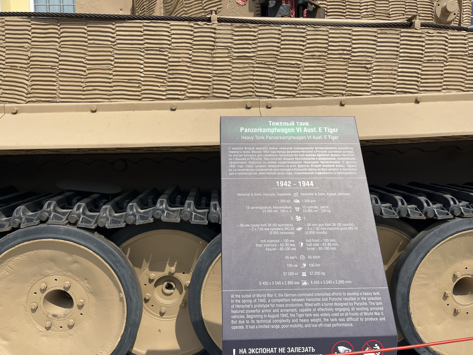The image shows a close-up of the tracks and road wheels of a German Panzerkampfwagen VI Ausf. E Tiger heavy tank, a museum exhibit at the Military Technology Museum in Verkhnyaya Pyshma, Russia. A descriptive plaque, with text in Russian and English, is positioned in front of the tank. The plaque details the tank's specifications and history, noting it was developed in 1942 and saw action on all fronts of World War II. The tank's camouflage exterior is visible, with a textured pattern and a thick rope running along the top. The environment appears to be outdoors, with sunlight casting shadows. No people are visible in the frame.