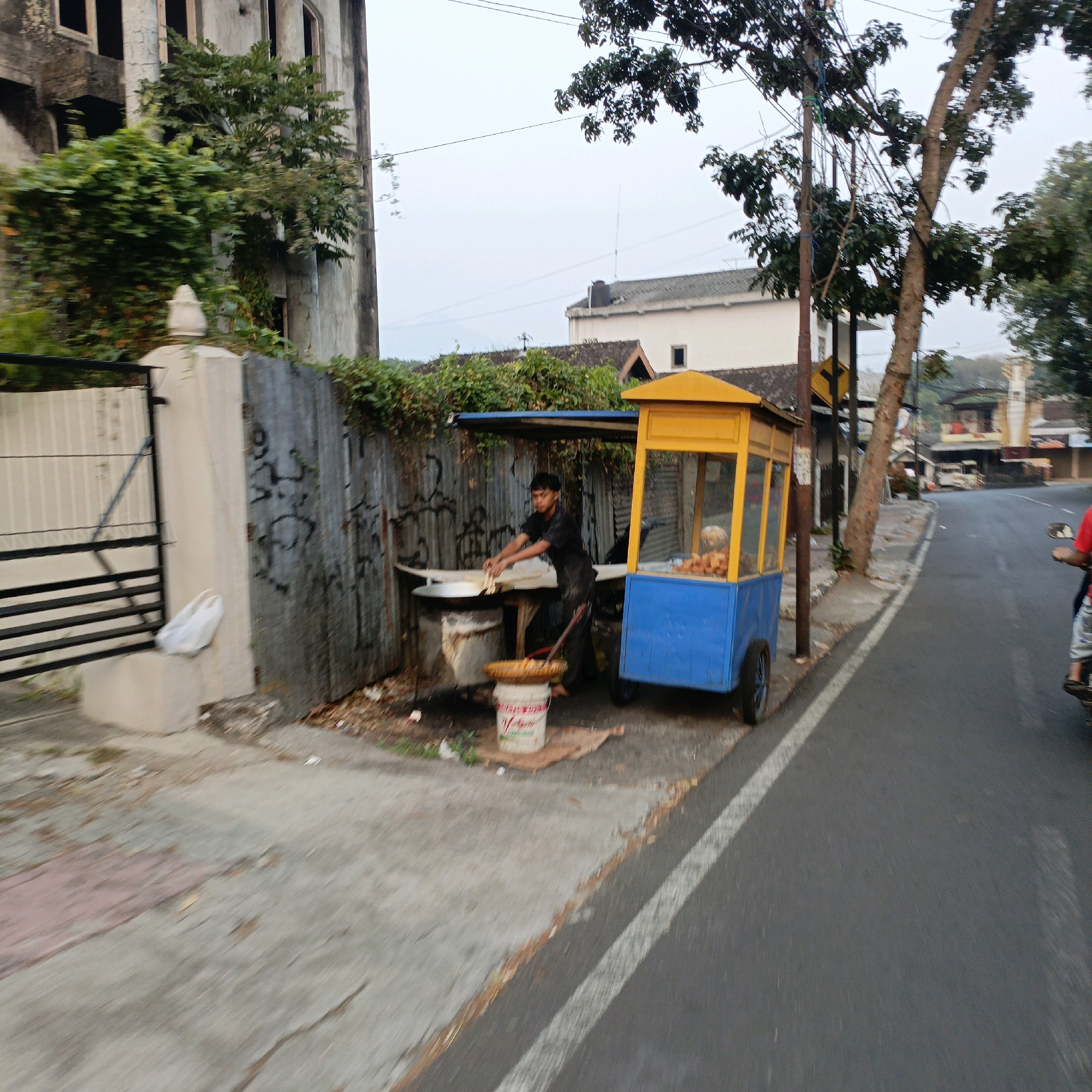 In Salatiga, Indonesia, a food vendor operates a blue and yellow cart on the side of a street. A young man, wearing a black t-shirt and dark pants, stands behind a corrugated metal stall, tending to a large metal wok or fryer. He is in the process of cooking or preparing food. In front of the stall, a white bucket with red lettering is placed on a piece of cardboard. The food cart is equipped with a glass display case filled with food items. The scene is set against a backdrop of a weathered building with overgrown greenery and a fence with graffiti. The street shows signs of wear, with some debris and foliage along the edges. A person on a motorcycle, wearing a red shirt, is visible in the right portion of the image, moving away from the vendor. The sky is overcast, suggesting a cloudy day.