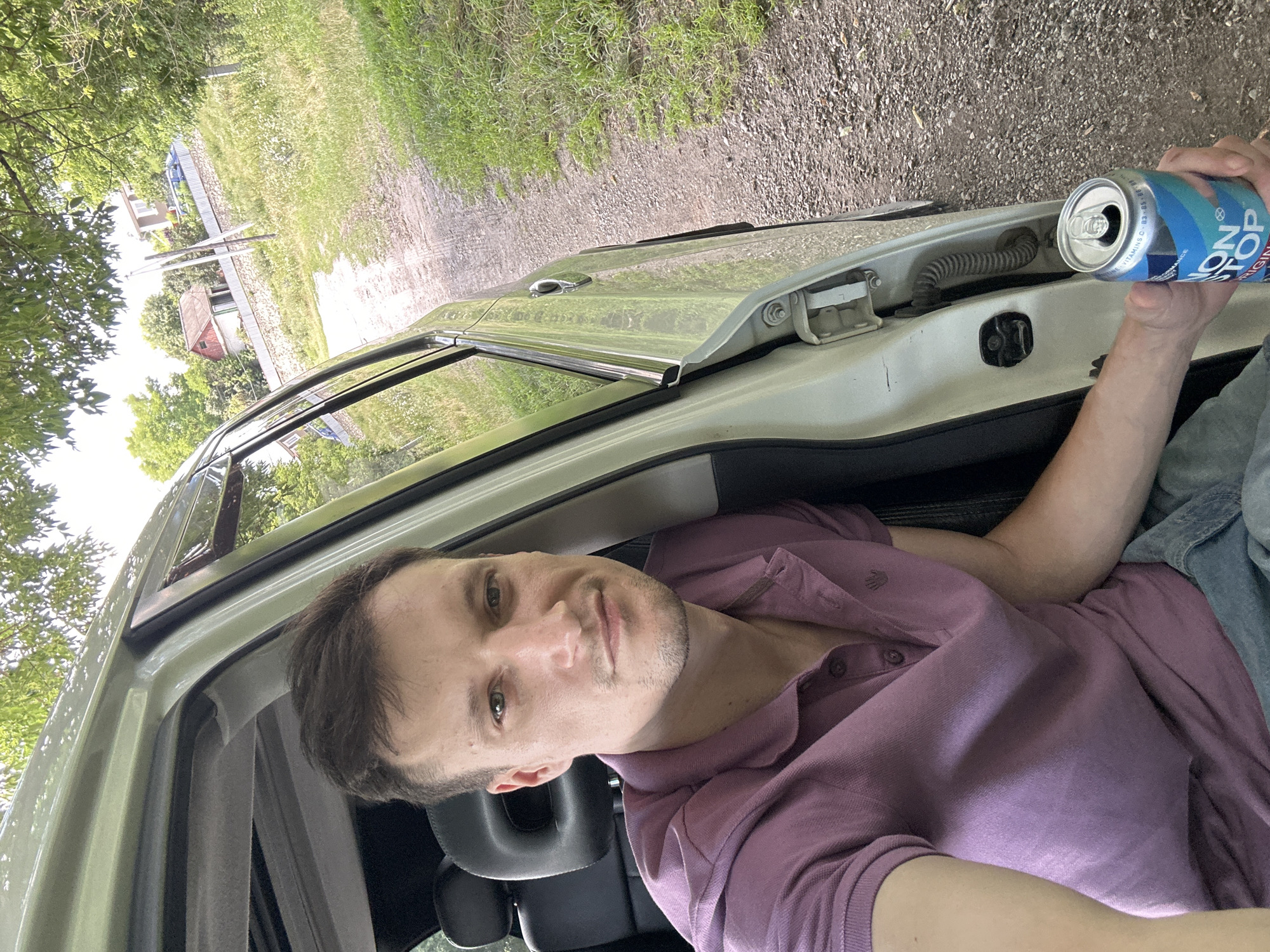 A man with short brown hair and a purple polo shirt is sitting in the driver's seat of a light-colored SUV. He is holding a can of "Non Stop" energy drink in his right hand. The car door is open, revealing a gravel road with green grass and trees on either side. In the background, there are houses and a fence. The scene appears to be a rural or suburban area. The image was taken in Makiv, Ukraine.