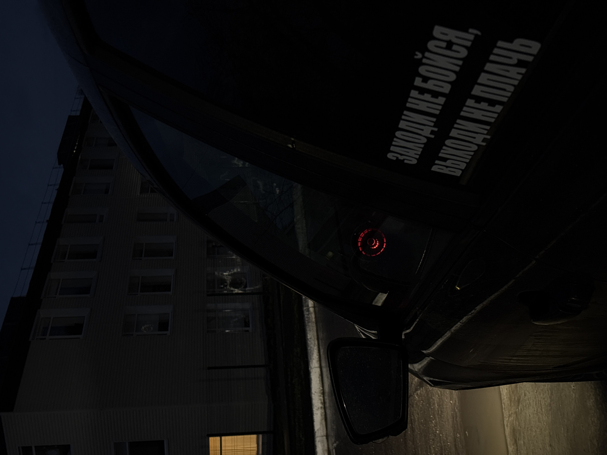 The image depicts a car parked on a street in Chaikovsky, Russia, at night. A multi-story apartment building is visible in the background, with some windows illuminated. The car features a sticker on its window with text written in Russian, which translates to "Come in, don't be afraid. Go out, don't cry". The car's side mirror is visible and wet, suggesting recent rain. The scene is dark and appears to be urban.