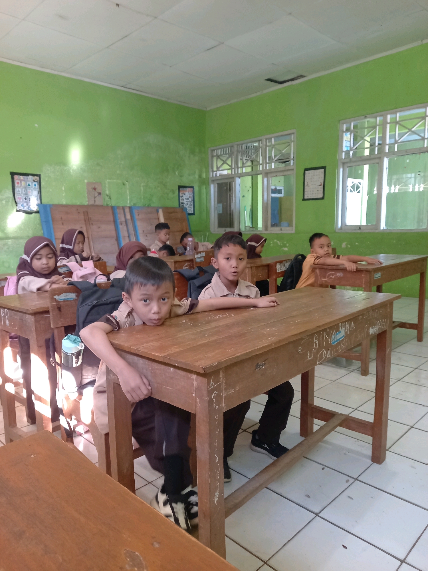 The image depicts a classroom scene in Kebaturan, Indonesia. Several young students, likely of elementary school age, are seated at wooden desks. They are dressed in school uniforms, which appear to be tan shirts and dark pants or skirts, with some girls wearing headscarves. The classroom has green walls and a tiled floor. Sunlight streams in from large windows on the right side of the frame, illuminating parts of the room and casting shadows. The desks are simple wooden structures, some with writing or graffiti on their surfaces. The students' expressions vary, with some looking towards the camera, others looking down at their desks, and some appearing a bit restless. The overall atmosphere suggests a typical school day.